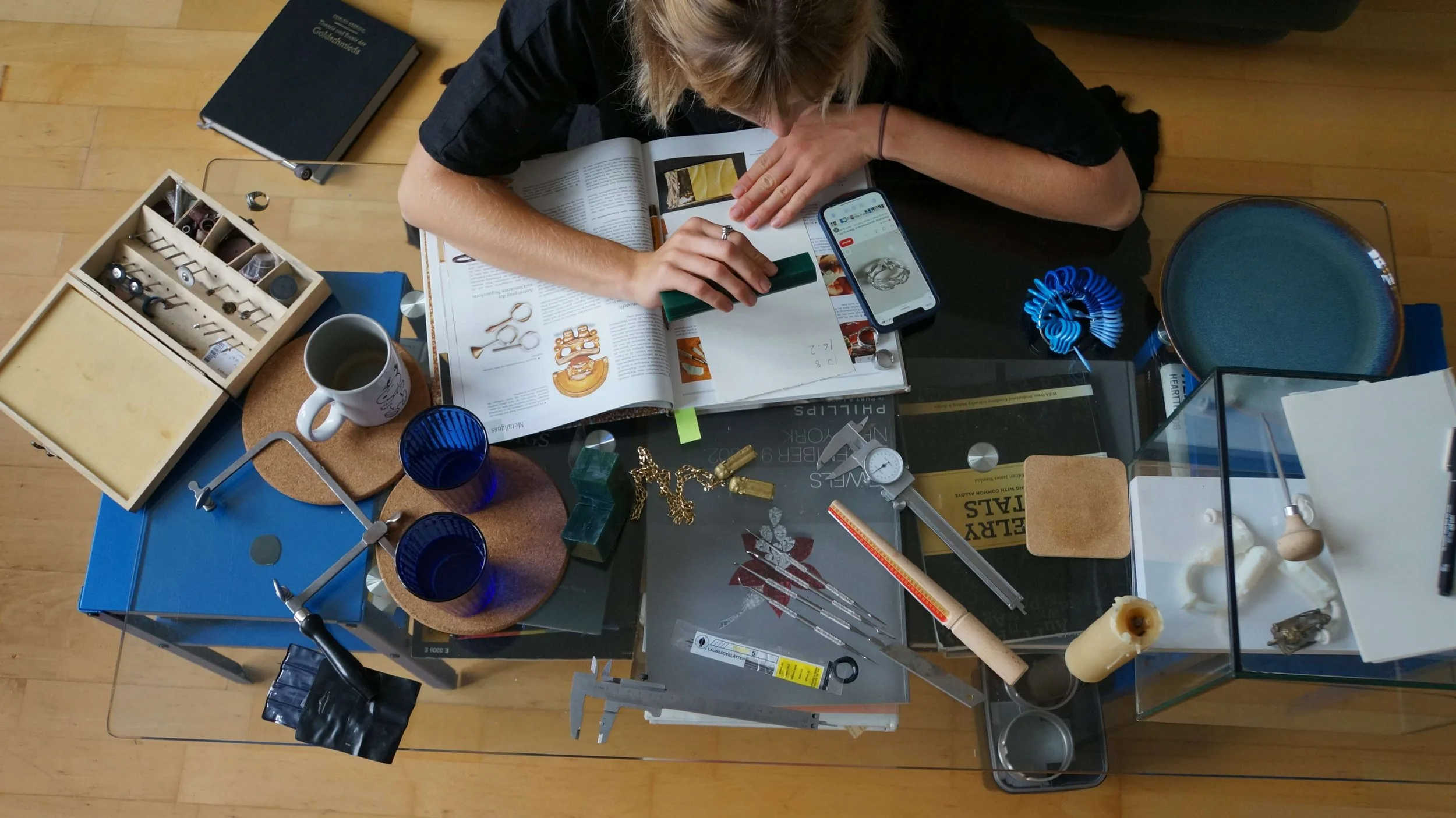 Overhead view of person at a cluttered glass table with jewelry, tools, a smartphone, and books.