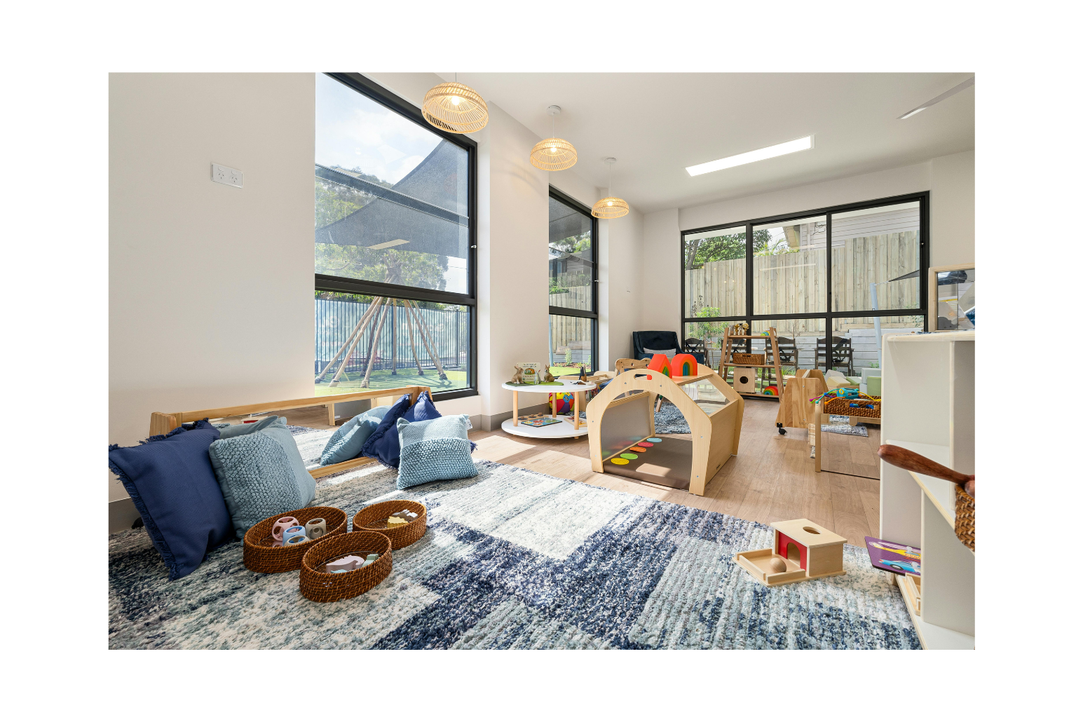 Bright and spacious indoor playroom with large windows, wooden flooring, and colorful children's toys, blankets, and cushions.