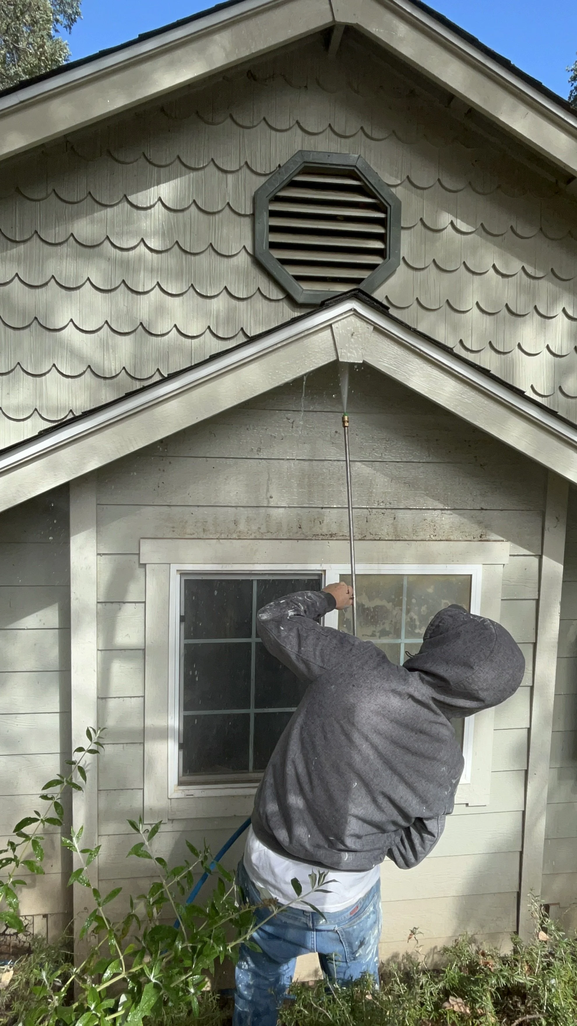 A person in a gray hoodie and blue jeans power washing a house.