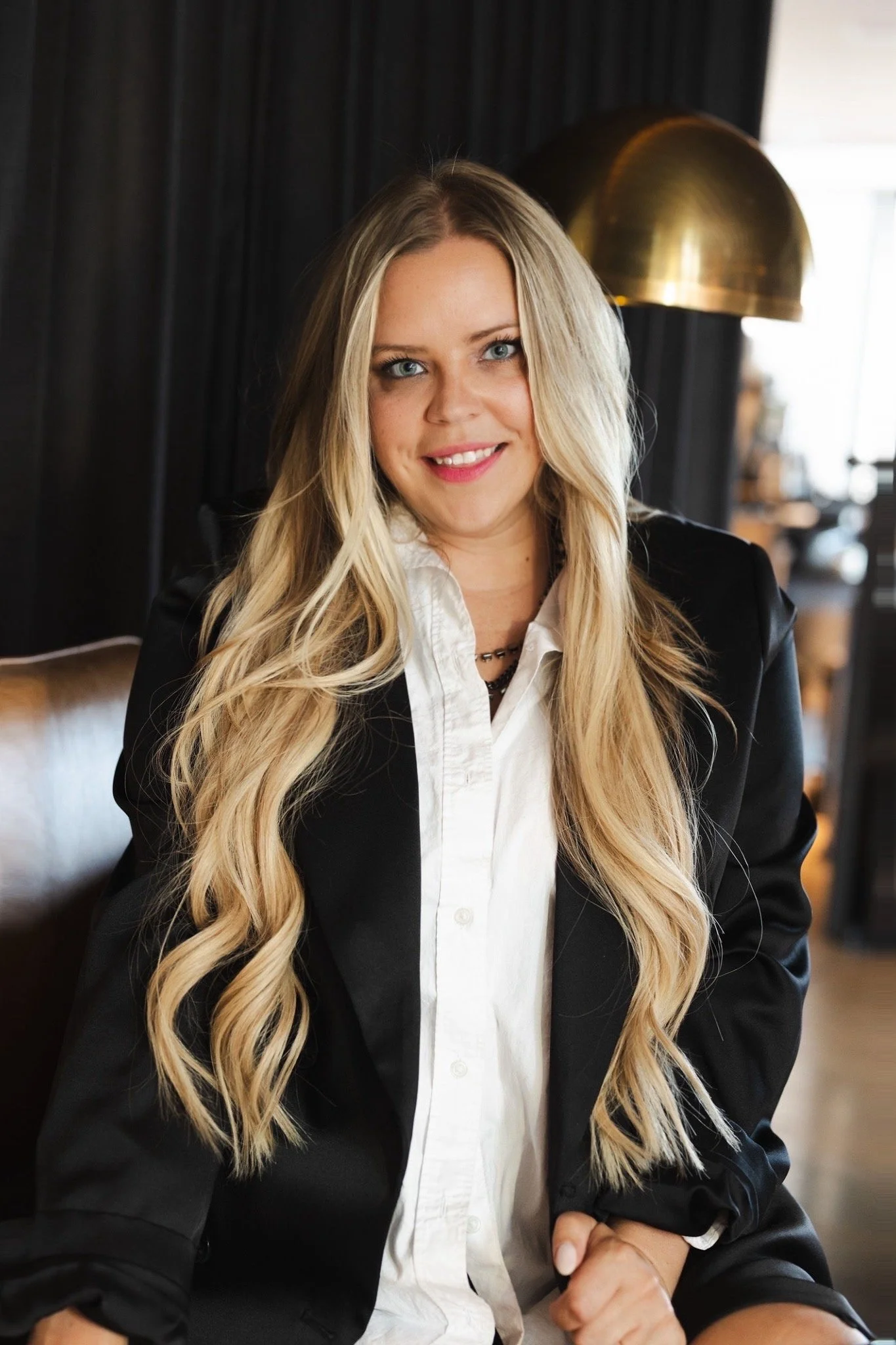 A woman with long blonde hair and blue eyes, wearing a white shirt and a black blazer, smiling indoors with dark drapes and a gold lamp in the background.