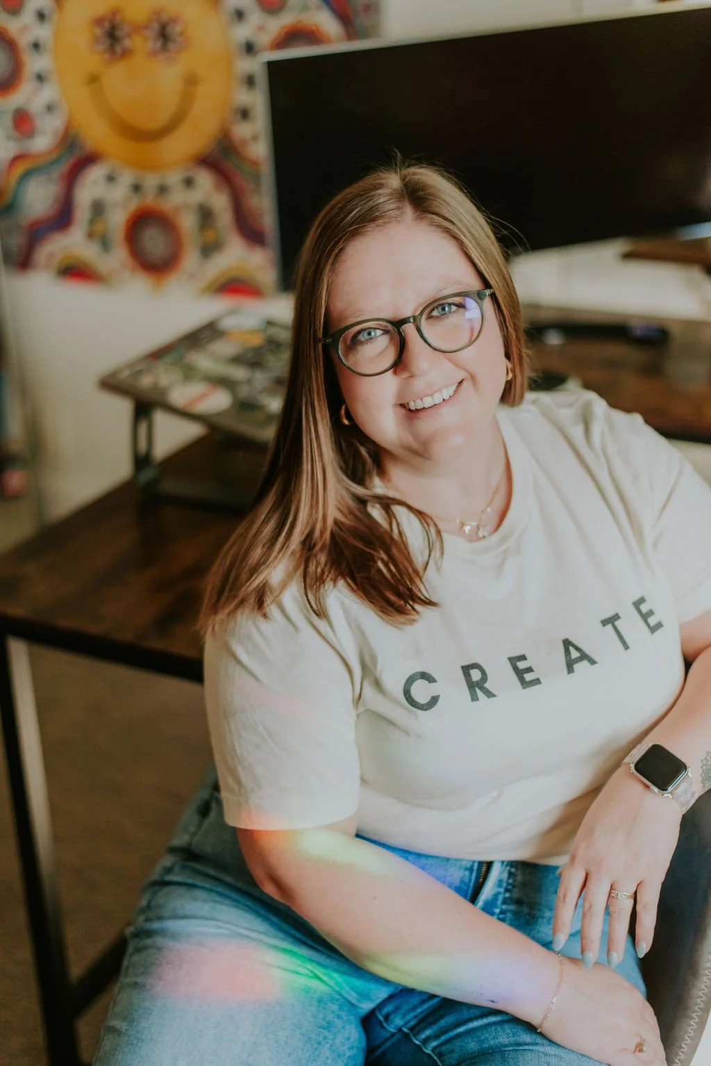 A woman with long brown hair, glasses, and a white T-shirt that says 'CREATE' sits at a desk, smiling at the camera. She is in a room with a colorful tie-dye wall and a computer monitor behind her.