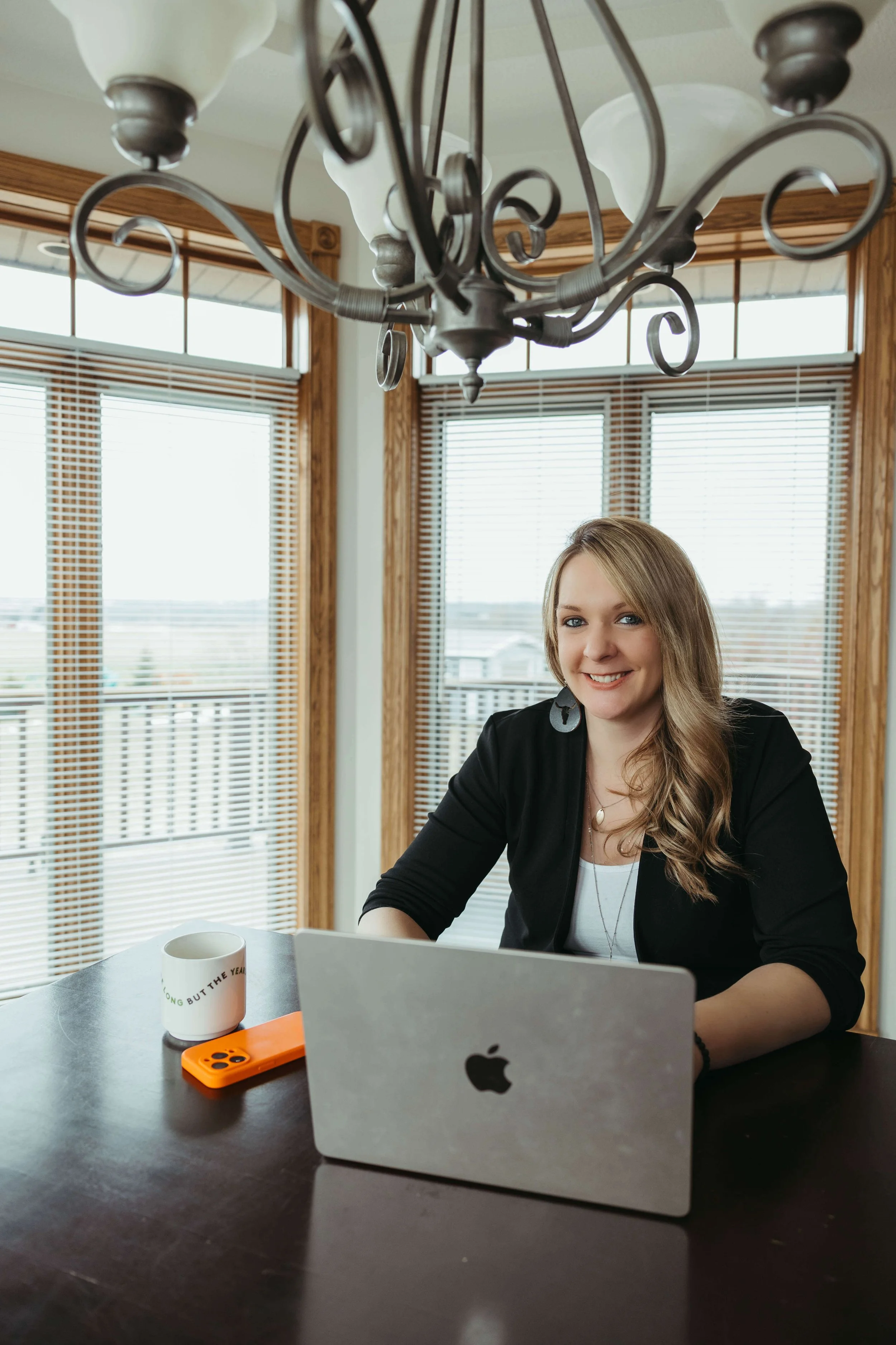 A woman sitting at a table with a laptop, coffee mug, and smartphone in a room with large windows and wooden trim, smiling at the camera.