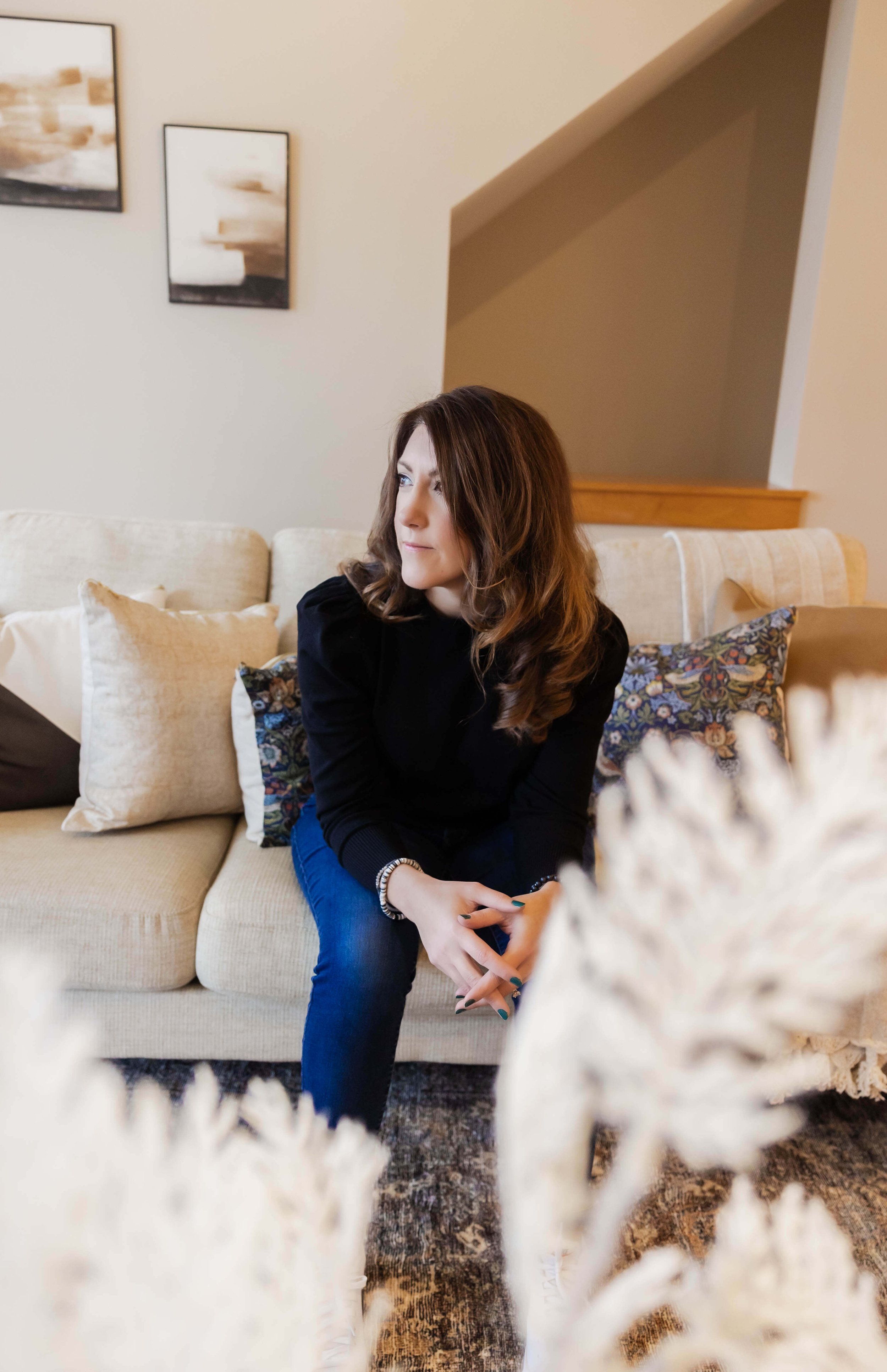 A woman with shoulder-length brown hair sits on a beige sofa with decorative pillows in a living room, gazing thoughtfully to her left.