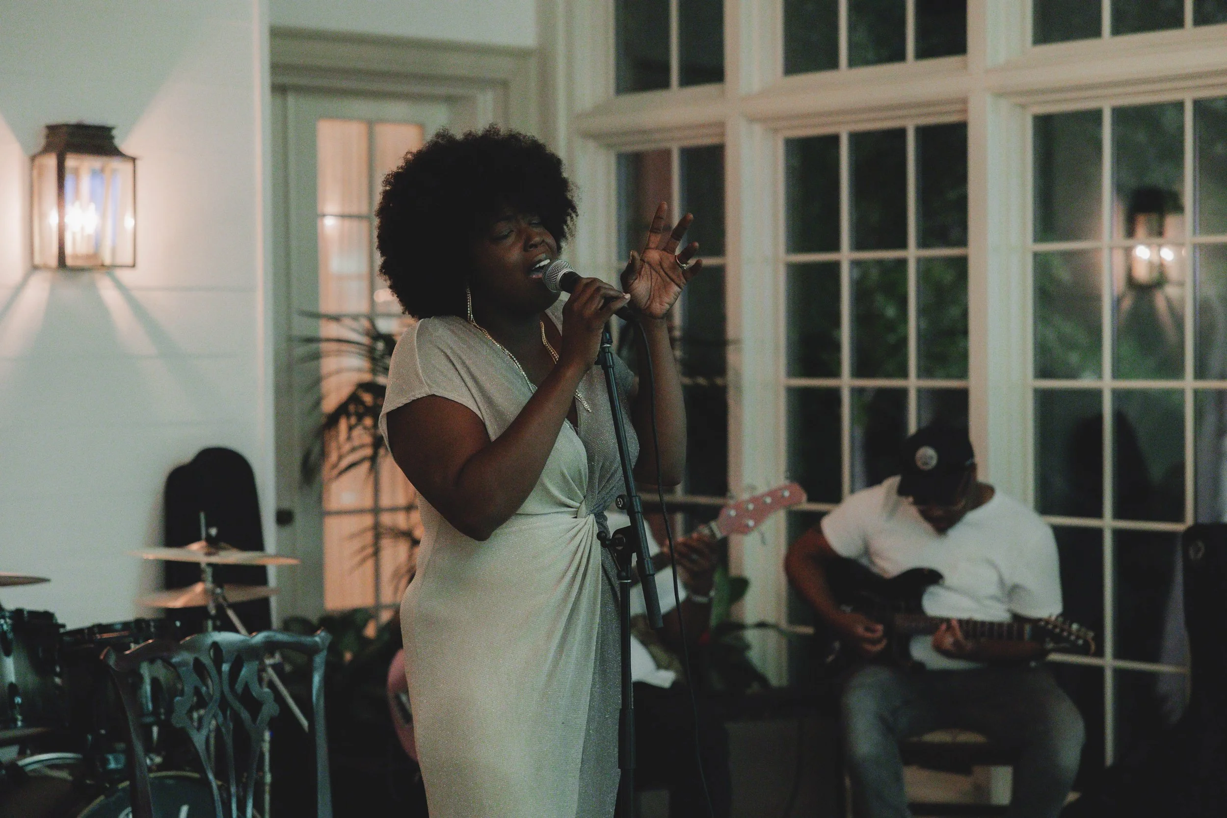 A woman singing into a microphone with her eyes closed, wearing a light-colored dress, while a musician playing a guitar is in the background inside a room with large windows.