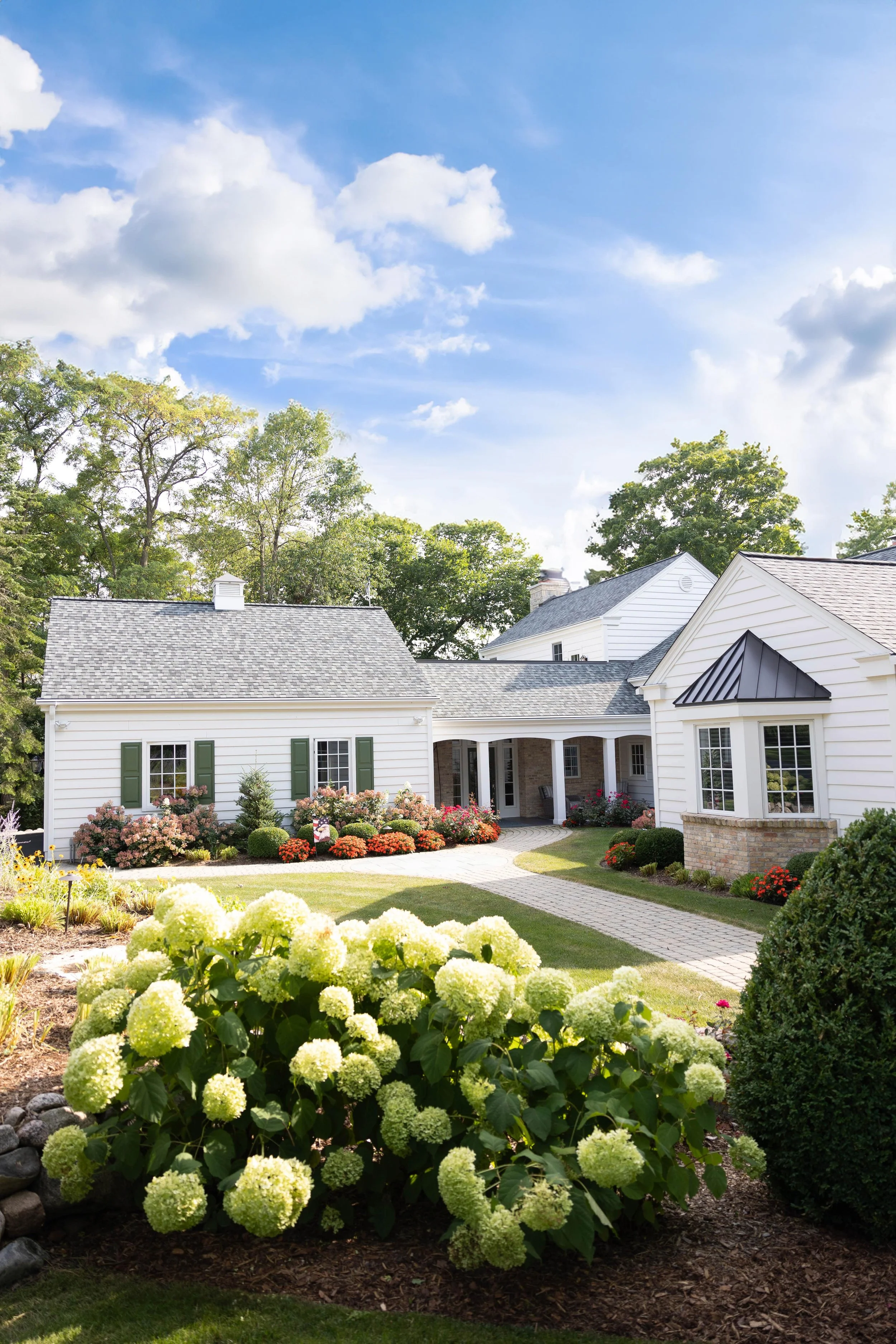 A well-maintained house with white siding, green shutters, a brick chimney, and a garden with colorful flowers and bushes under a partly cloudy sky.
