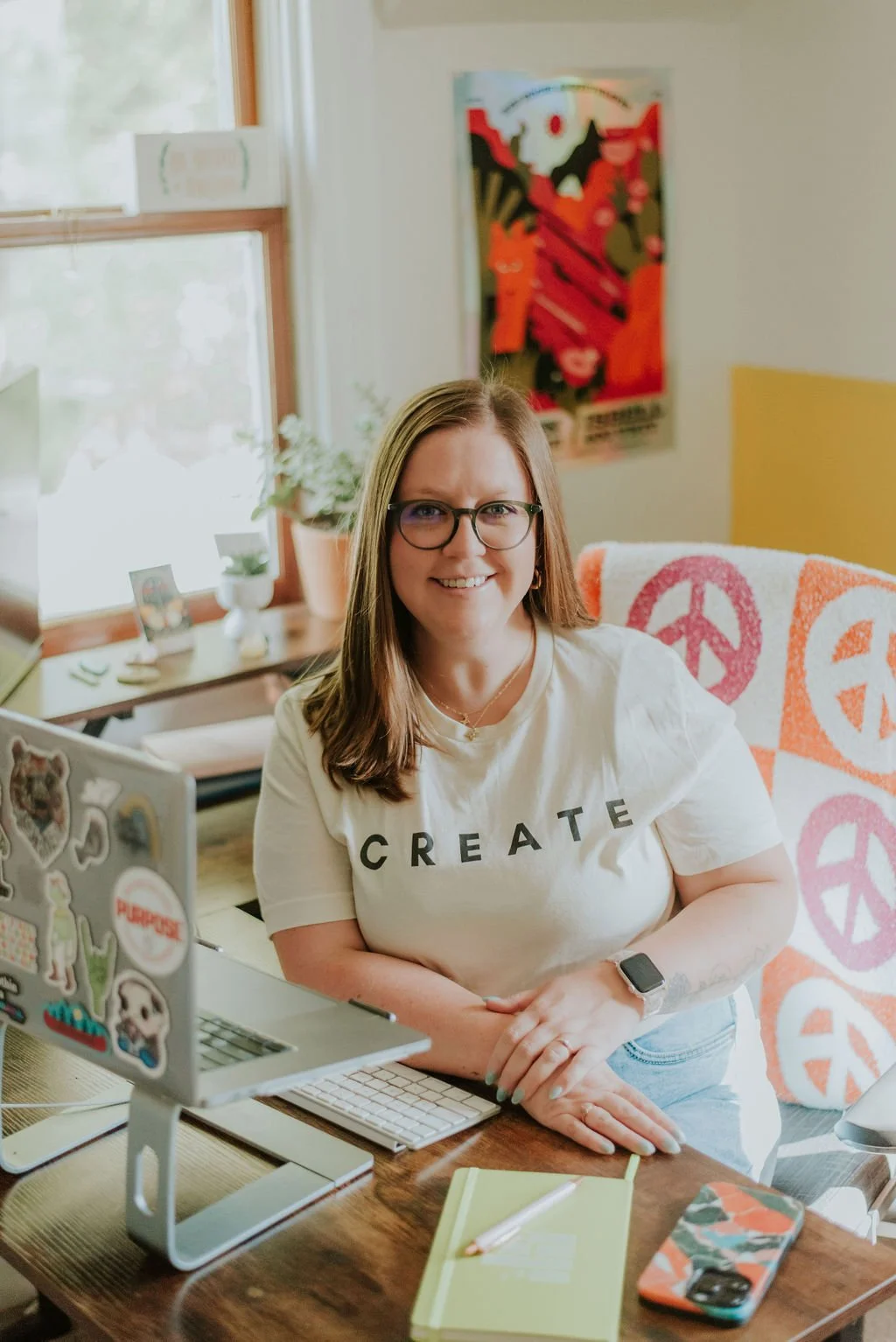 A woman with glasses and shoulder-length brown hair smiling at her desk. She is wearing a white t-shirt with the word 'CREATE' and sitting in a room with a window, a colorful peace sign blanket, and a poster on the wall.