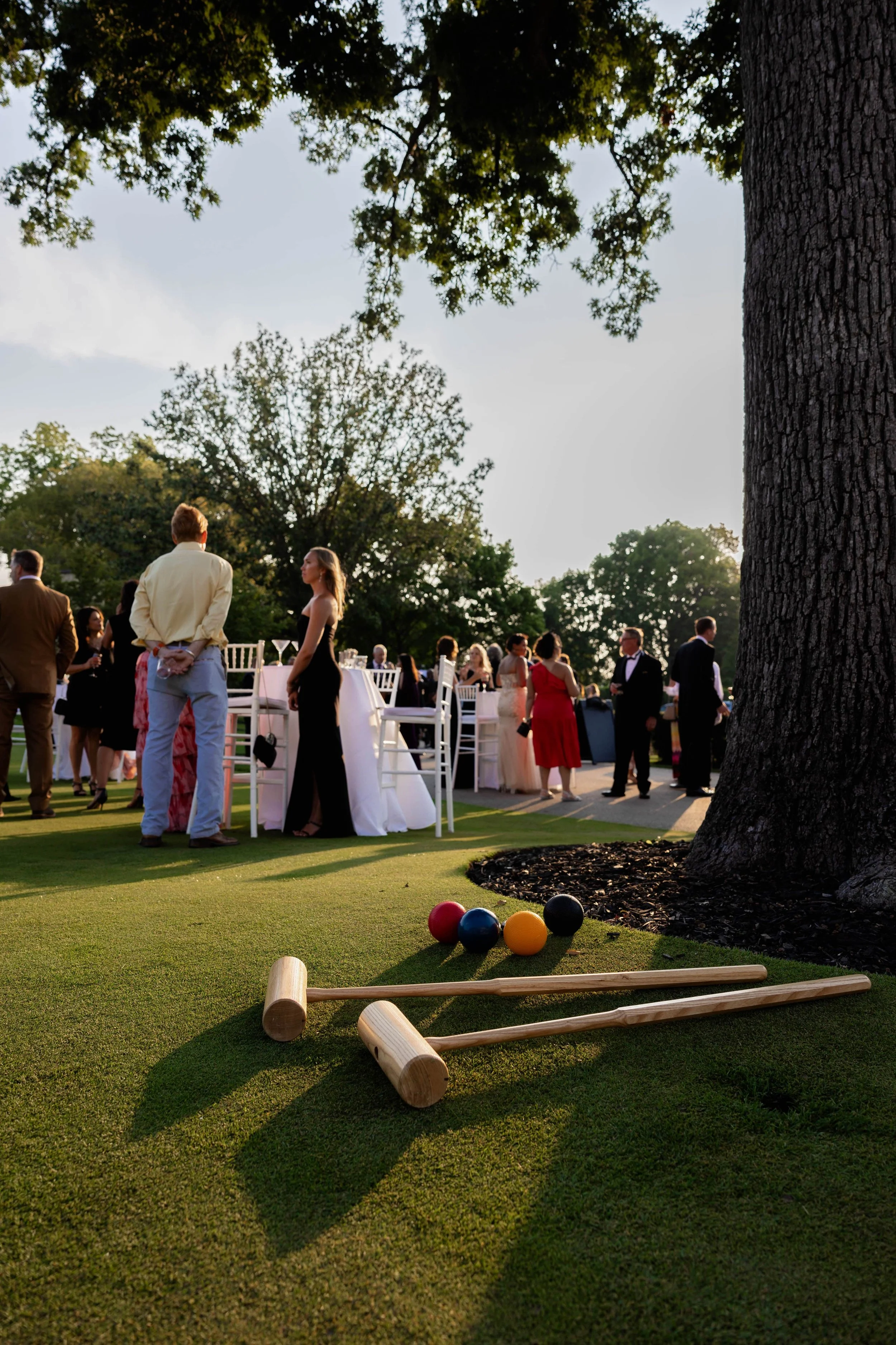 Outdoor gathering with people in formal attire, round tables, and chairs on a grassy area with trees, lawn game equipment in the foreground, and sunlight filtering through the trees.