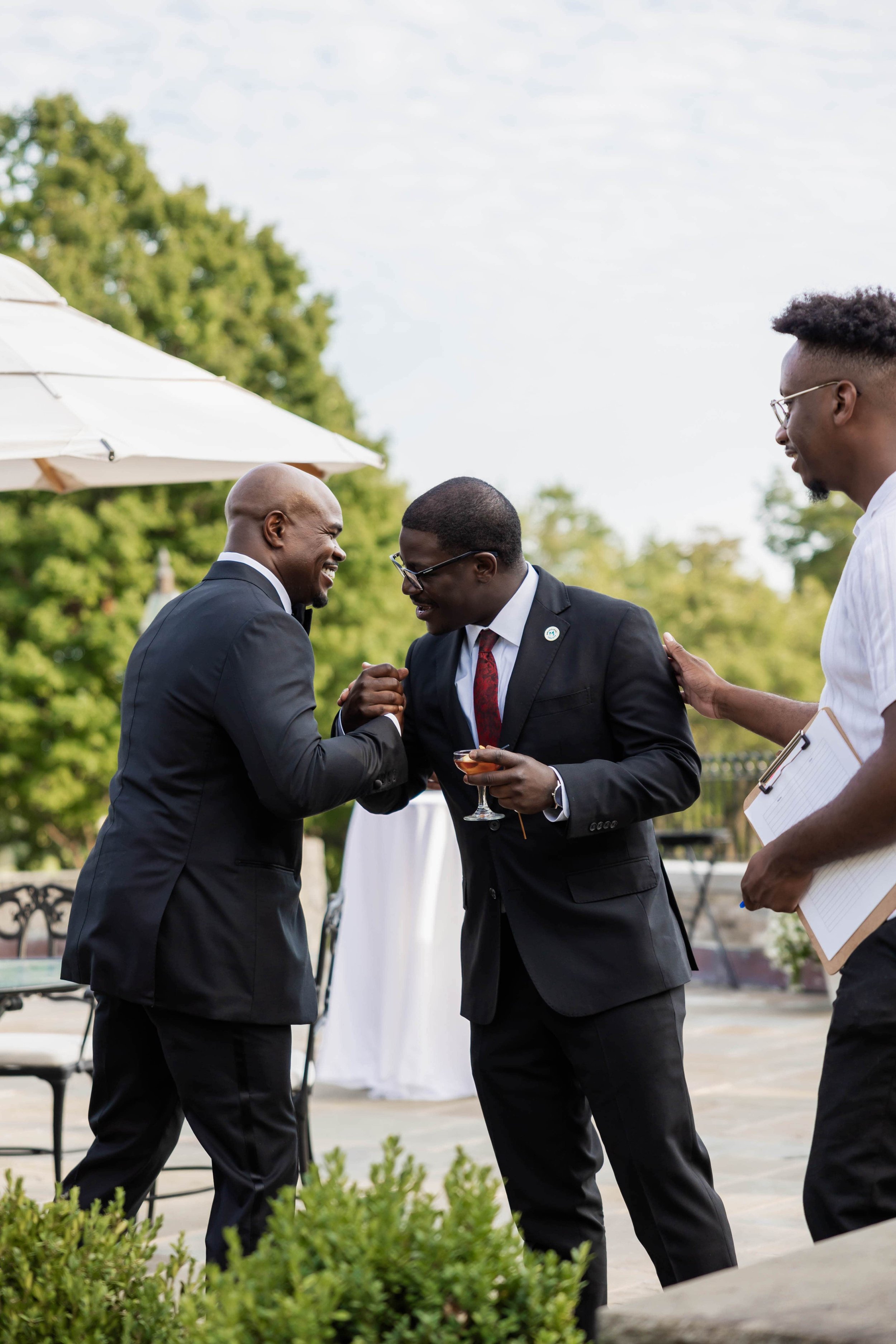 Three men in suits are at an outdoor event, engaged in a friendly arm-wrestling match while holding drinks and smiling, with greenery and an umbrella in the background.