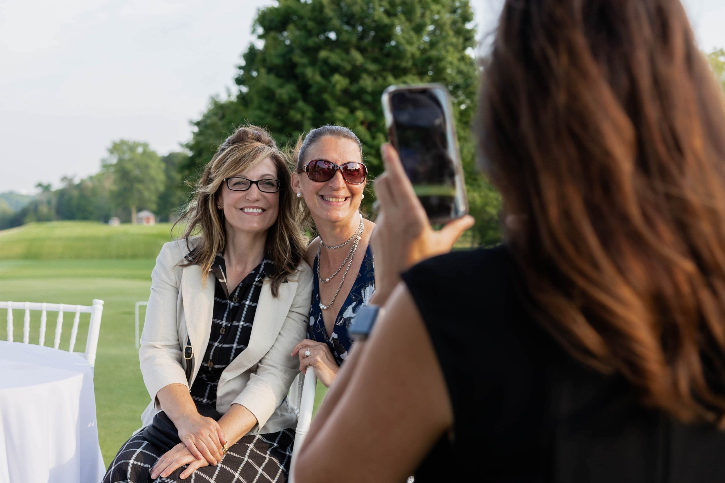 Three women outdoors, two seated and smiling at camera, one standing and taking a photo with a smartphone. Green trees and grass in background.