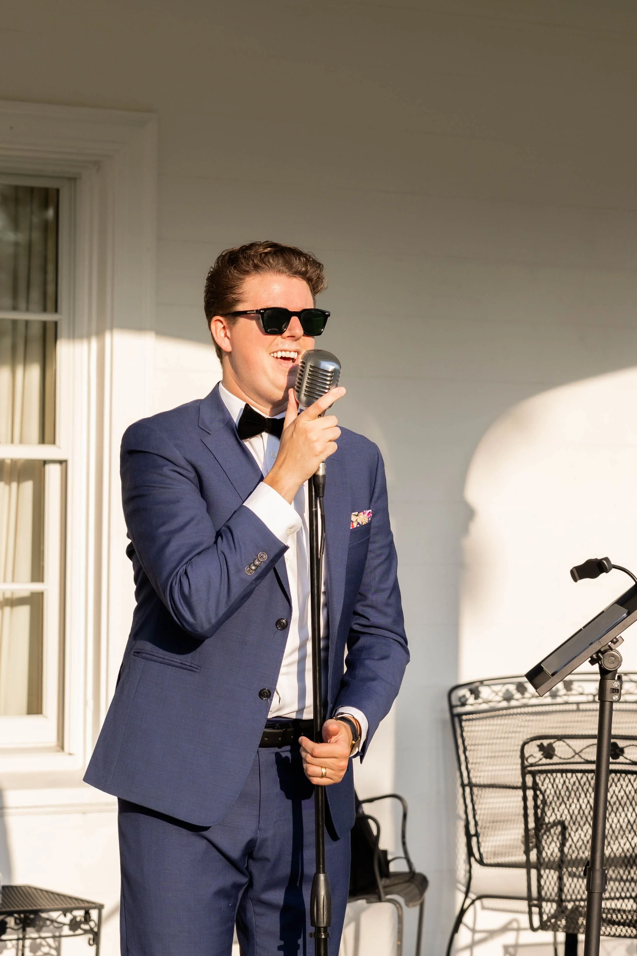 A man dressed in a blue suit, white shirt, and black bow tie, singing into a microphone on an outdoor porch, wearing sunglasses.