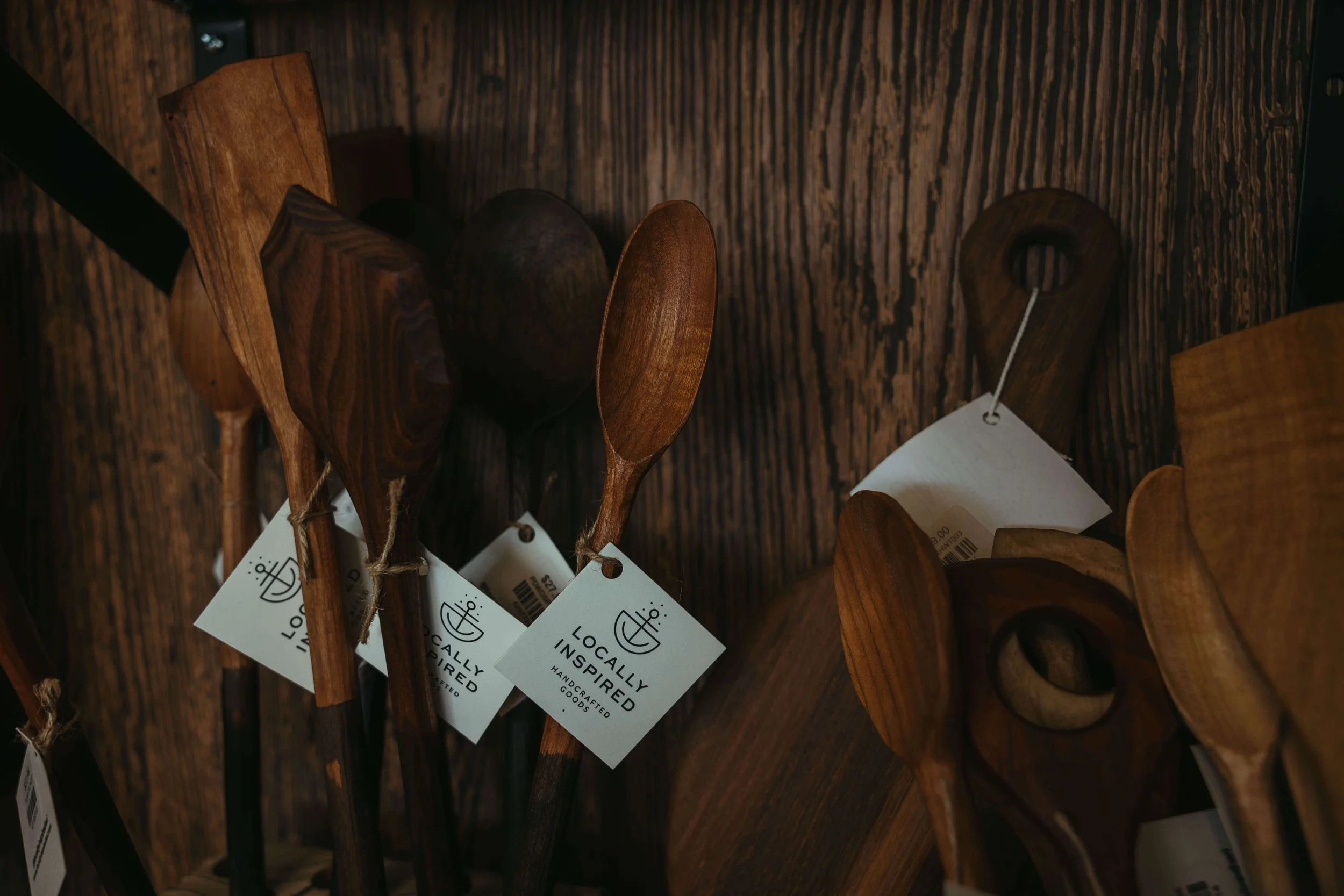 Set of wooden spoons and kitchen utensils hanging on a rustic wooden wall with tags reading "Locally Inspired".