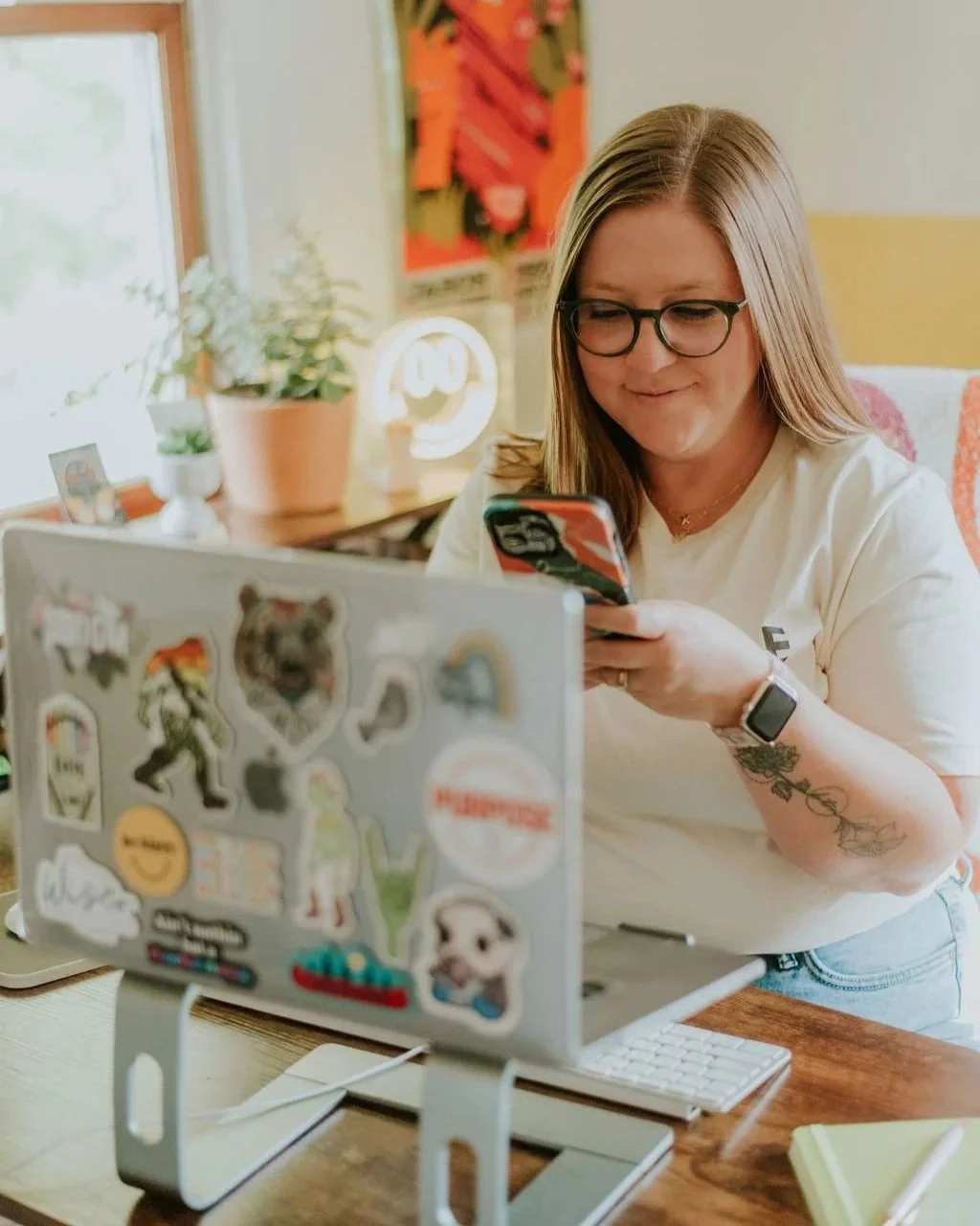 A woman with glasses and tattoos looks at her phone while working on a laptop decorated with stickers, sitting at a wooden table in a cozy room with plants and colorful posters.