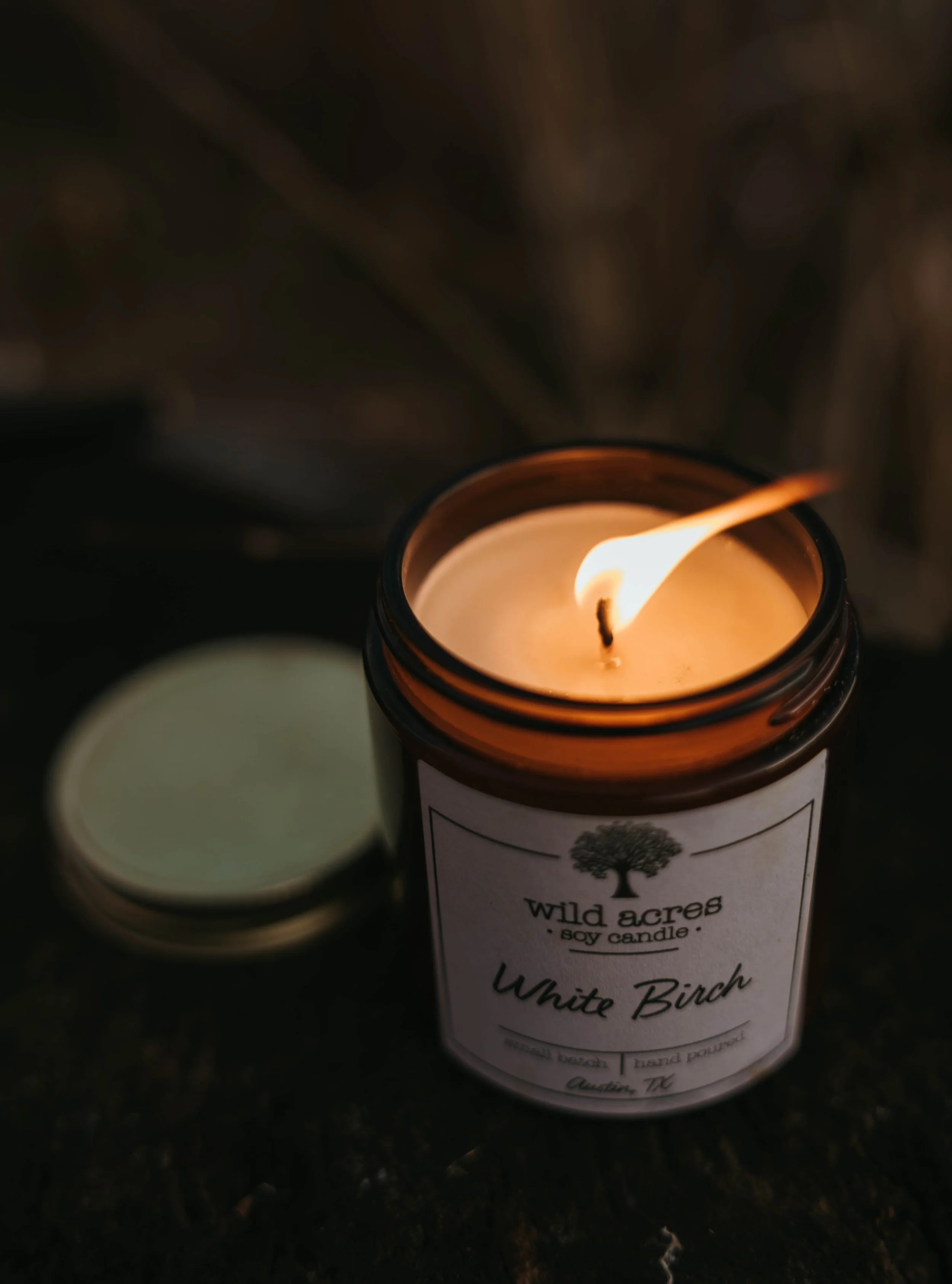 A lit white birch-scented candle in a brown jar with a white label, placed outdoors on a dark surface