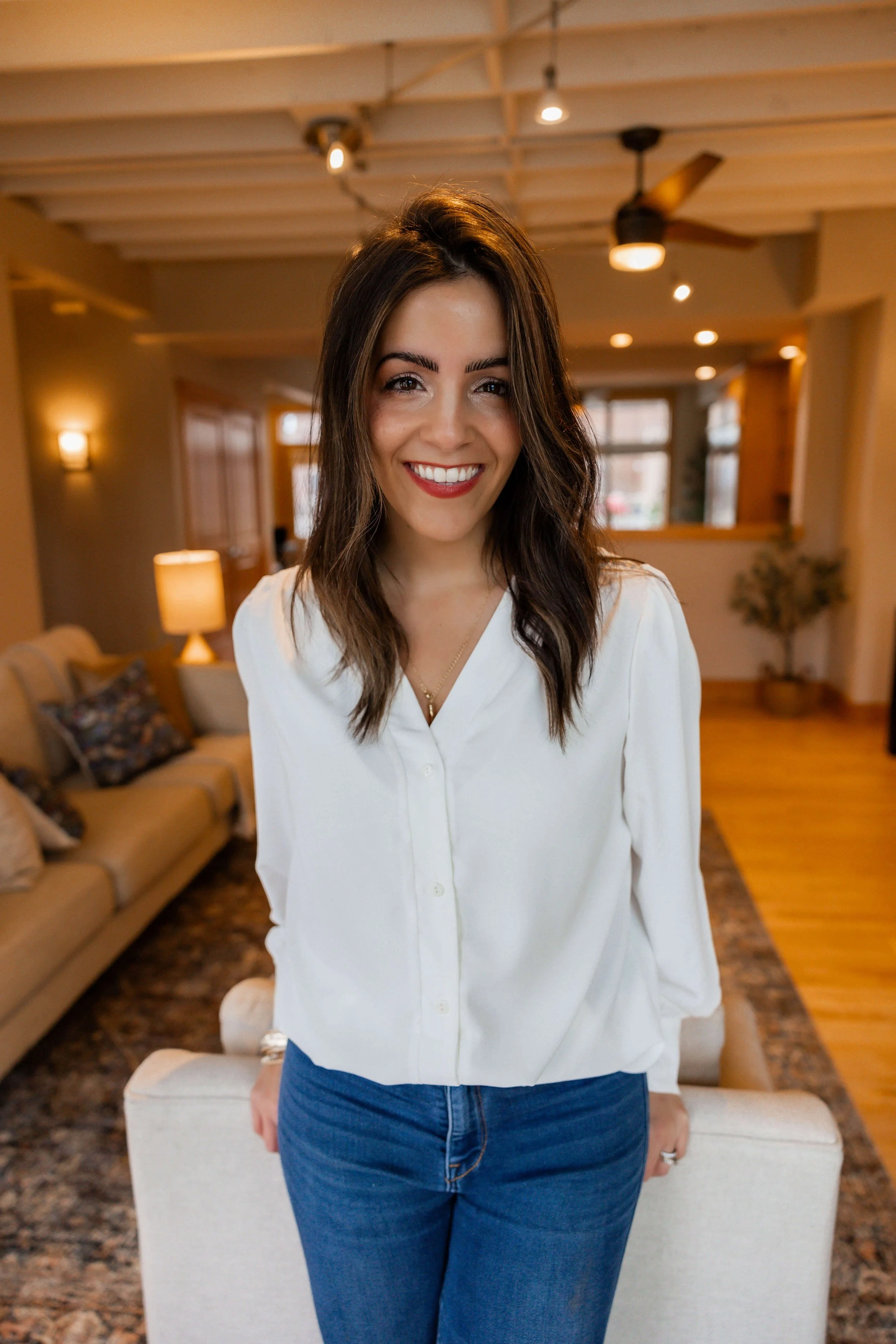 A smiling woman with medium-length brown hair, wearing a white blouse and blue jeans, standing in a cozy living room with beige furniture, a patterned rug, a lamp, and a ceiling fan.