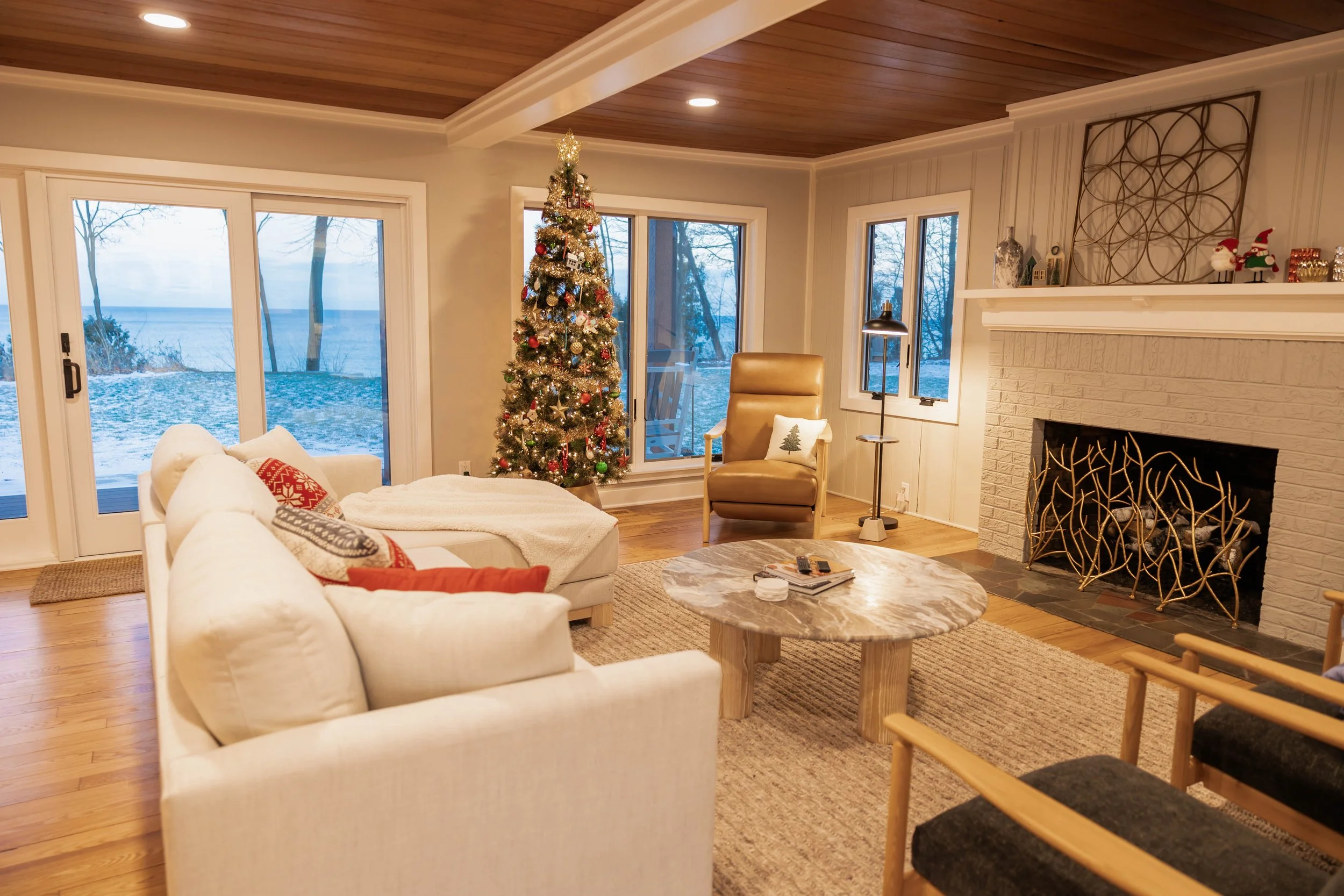 Living room with a decorated Christmas tree, white sofa, coffee table, beige armchair, and a fireplace with a decorative metal screen, overlooking a snowy outdoor landscape through large windows and glass doors.