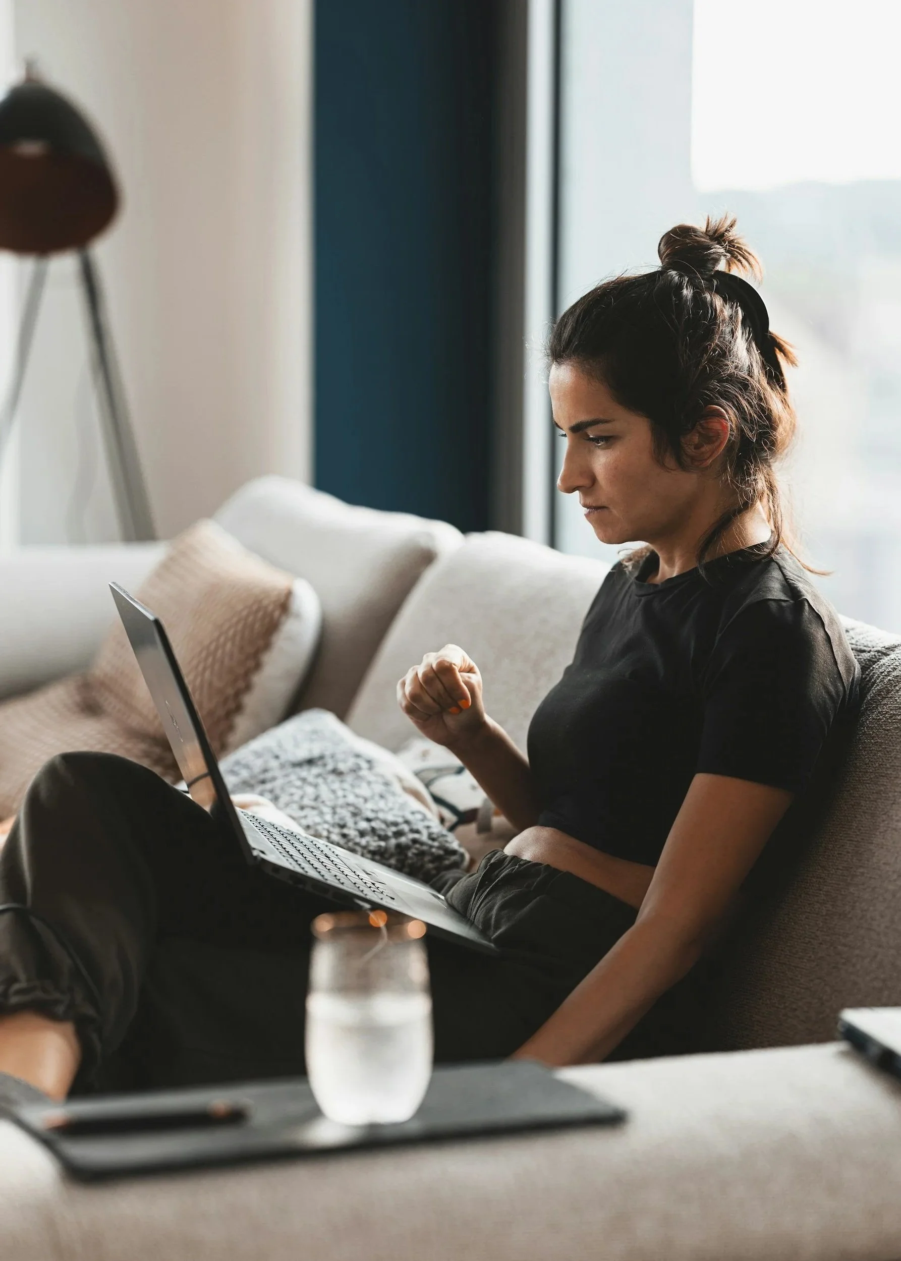 Woman sitting on a couch using a laptop, with a glass of water and a tablet on a table in front of her.