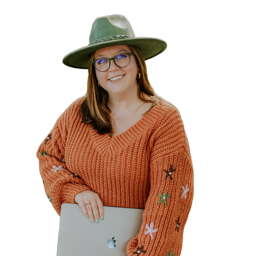 A woman smiling, wearing glasses, a wide-brimmed hat, and a knitted orange sweater with floral embroidery, holding a silver laptop.