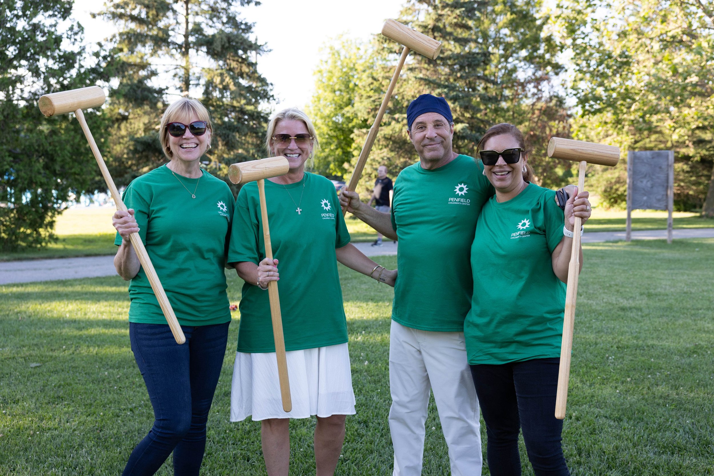Four adults standing outdoors on grass, smiling, holding wooden mallets, wearing green shirts with a logo, in a park with trees in the background.