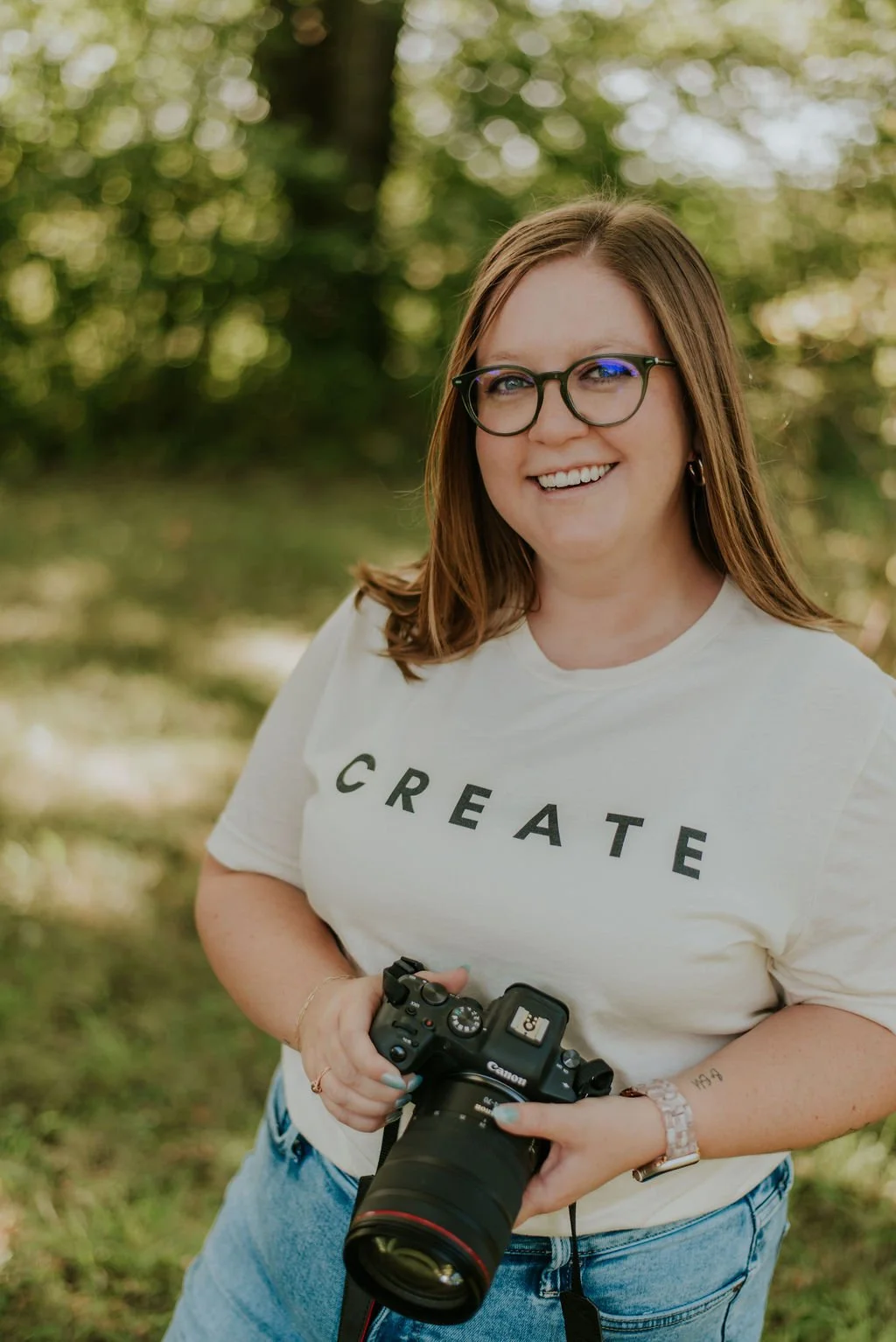 A smiling woman with glasses holding a professional camera outdoors, standing in a park or forested area.