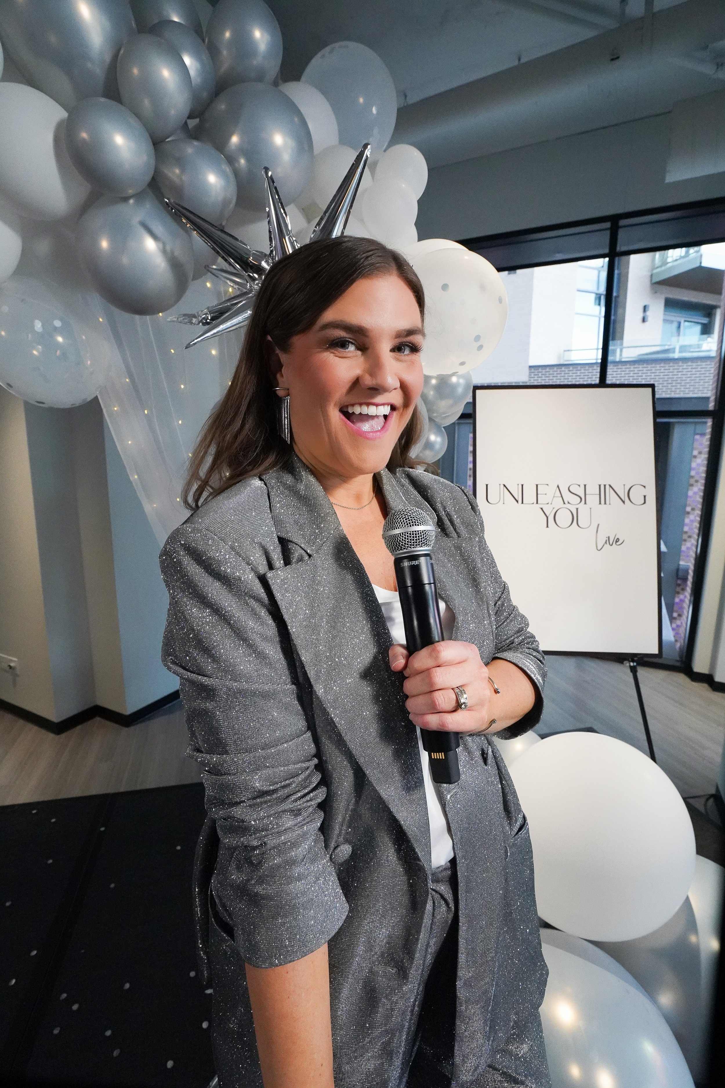 A woman in a sparkly silver blazer holding a microphone, smiling at a lively event with balloons and a sign that reads 'Unleashing You Live' behind her.