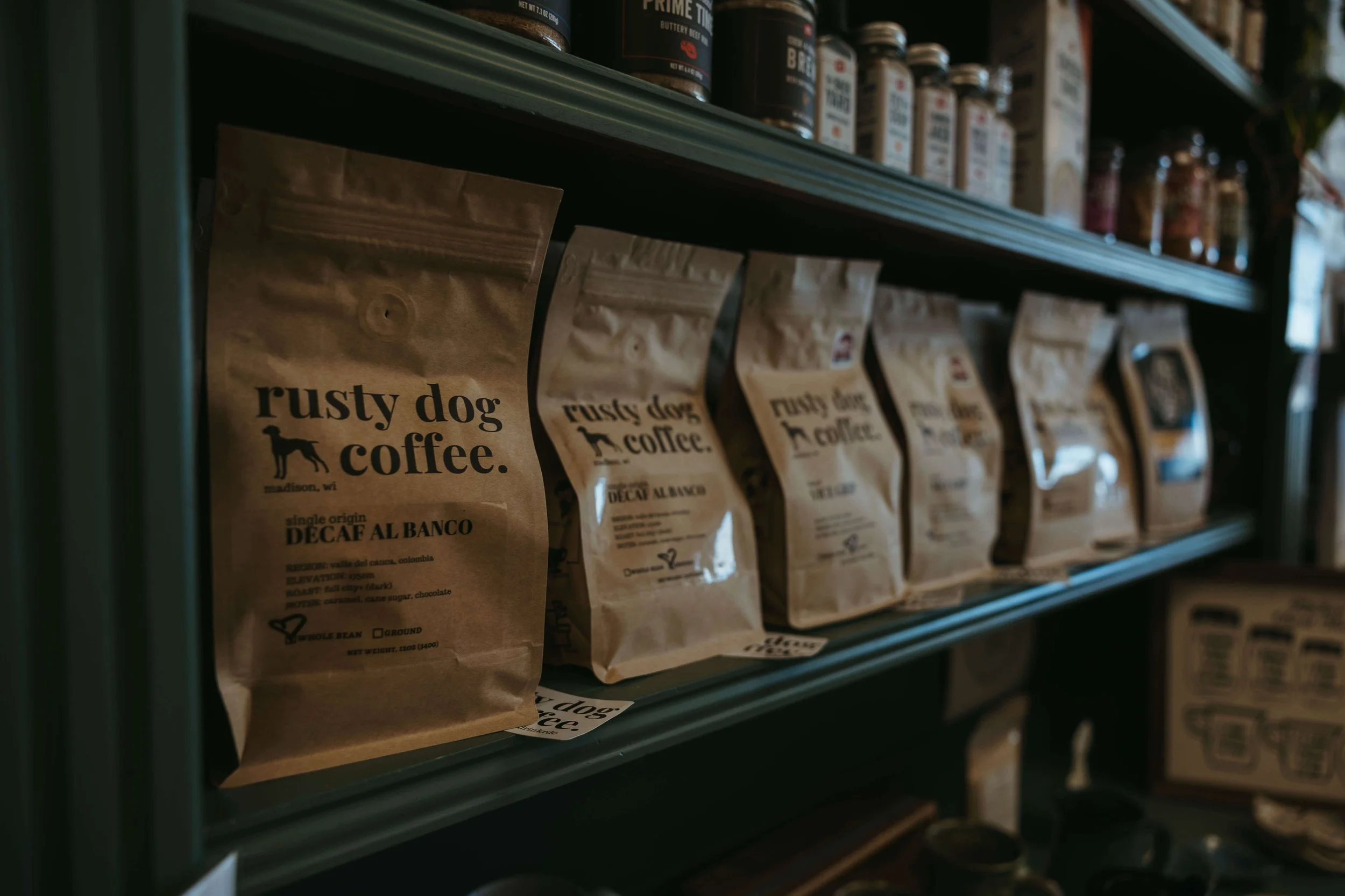 Shelf with brown paper bags of coffee from Rusty Dog Coffee, including Decaf Al Banco, and various jars and bottles on the top shelf.