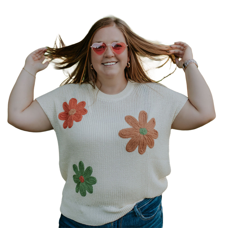 A smiling woman with red hair and pink sunglasses outdoors, adjusting her hair, wearing a white sweater with colorful felt flowers, standing in a park with green trees.