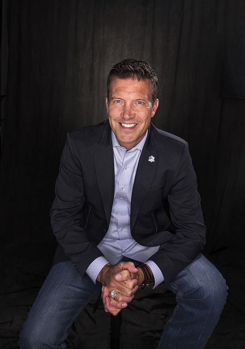A man in a dark blazer and jeans, sitting on a stool against a dark background, smiling and clasping his hands.