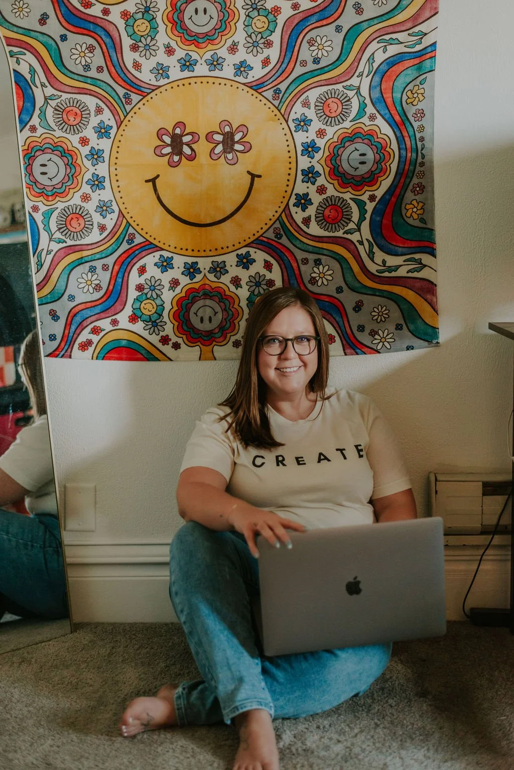 A woman with glasses sitting on the floor with a laptop on her lap, smiling. Behind her is a colorful tapestry with a large smiling yellow face, surrounded by smaller smiling faces, flowers, and rainbow-like patterns.