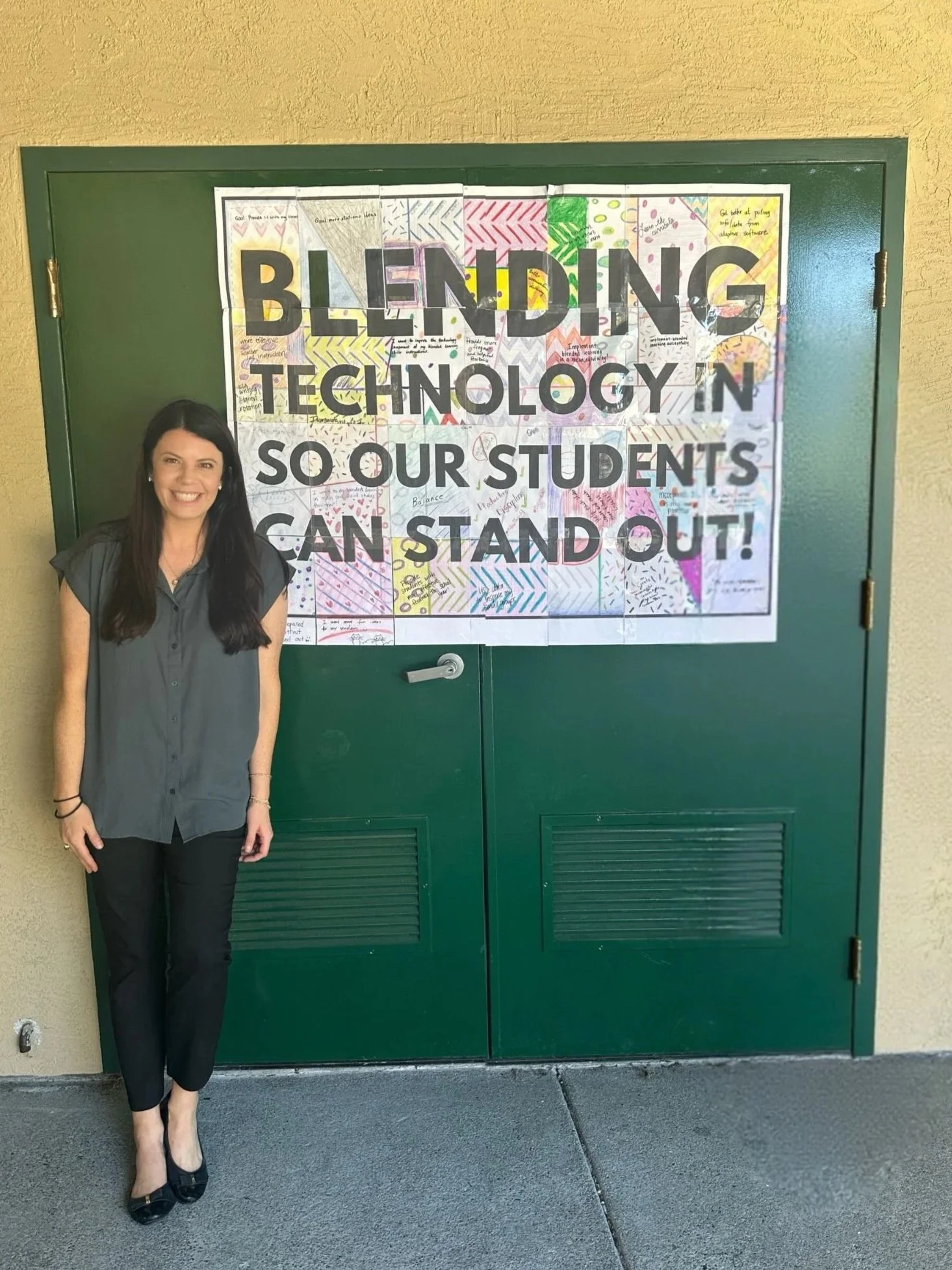 A woman standing beside a large banner on a green door with the words 'Blending Technology in So Our Students Can Stand Out' written on it.