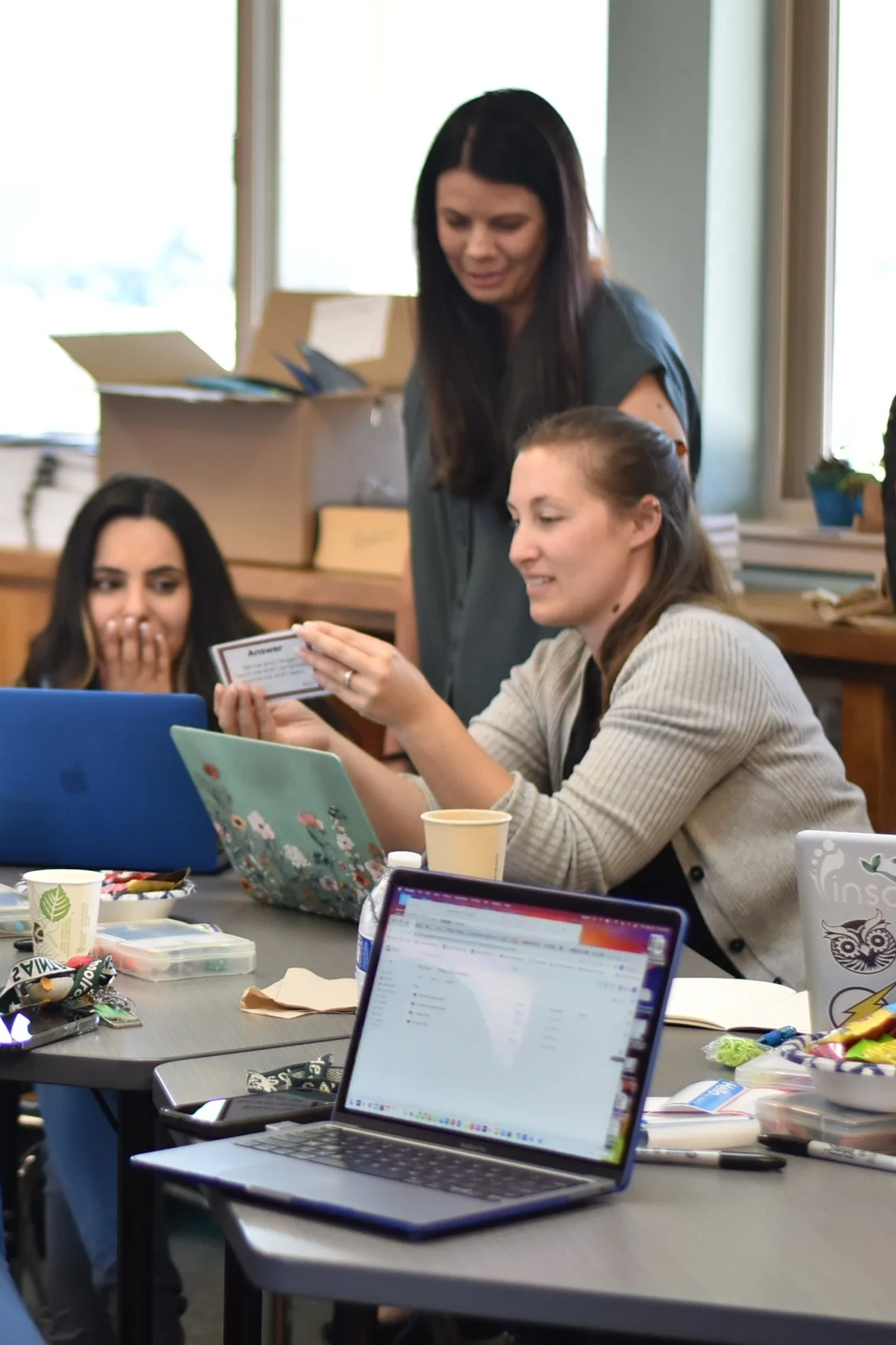 Three women sitting at a cluttered table with laptops, paper, and snacks, with one woman standing and handing a card to one of the seated women in a room with large windows.