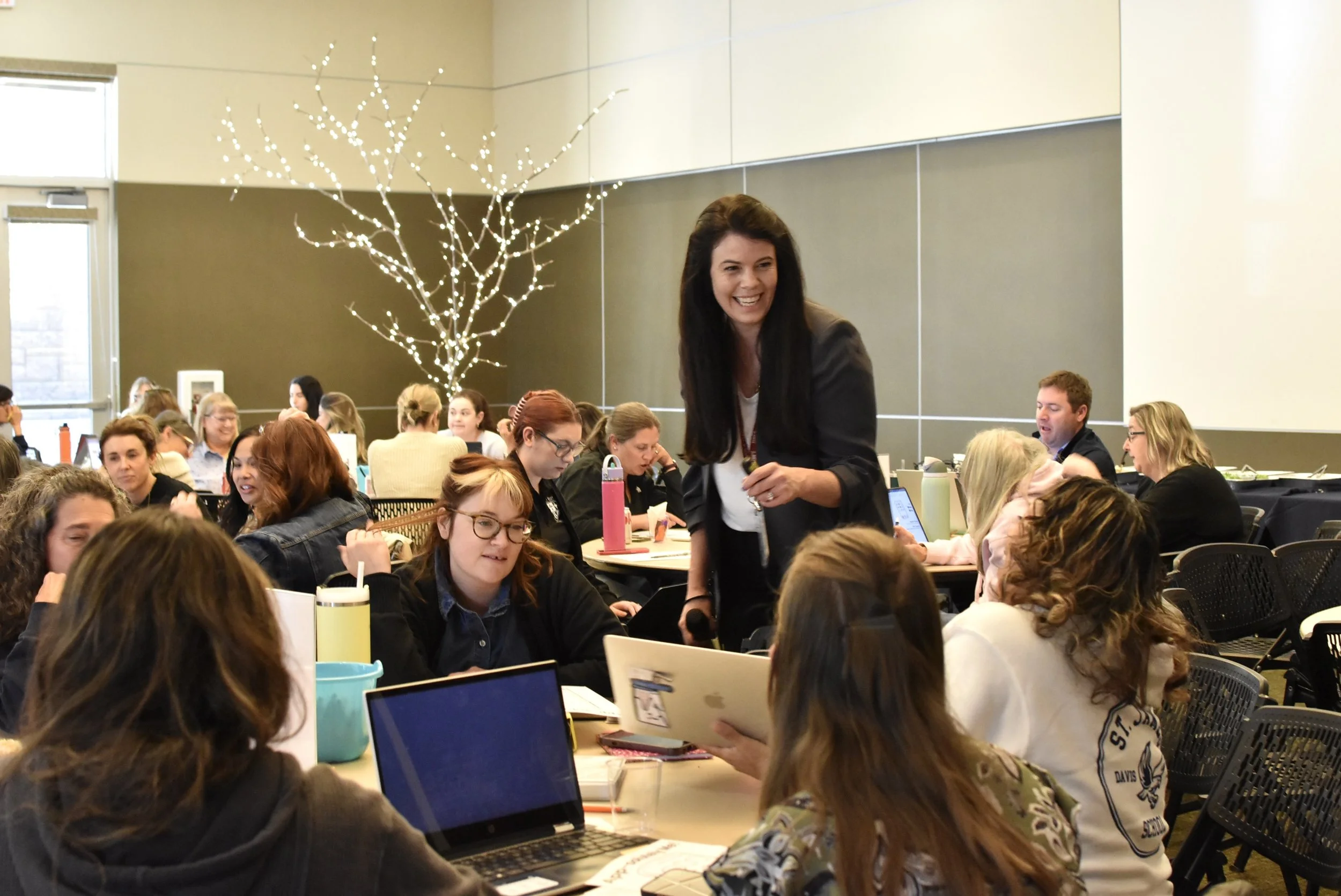 A woman standing and smiling in a conference room filled with people working on laptops, with a decorative illuminated tree in the background.