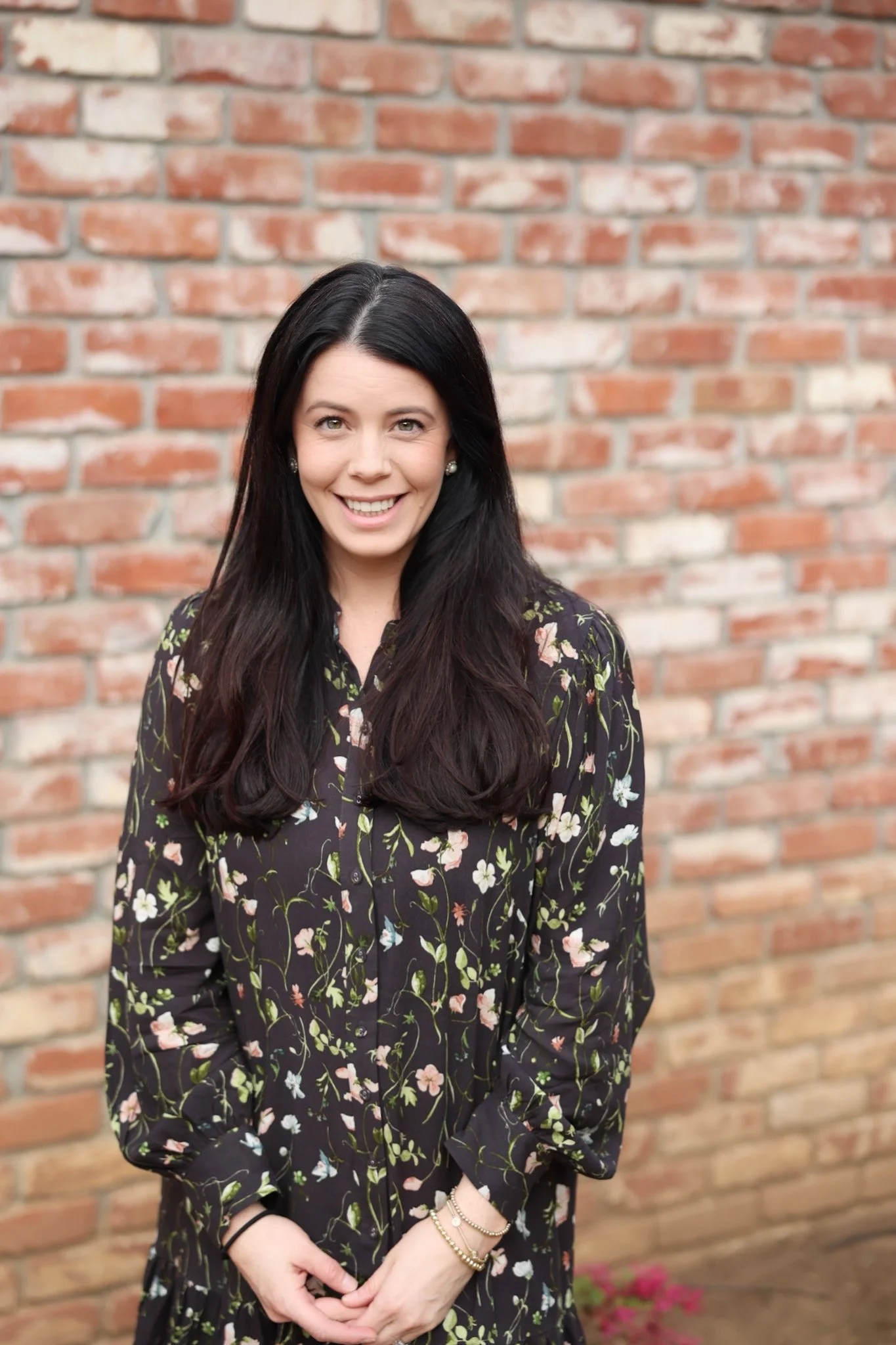 A woman with long dark hair, wearing a black floral dress, stands in front of a brick wall, smiling at the camera.