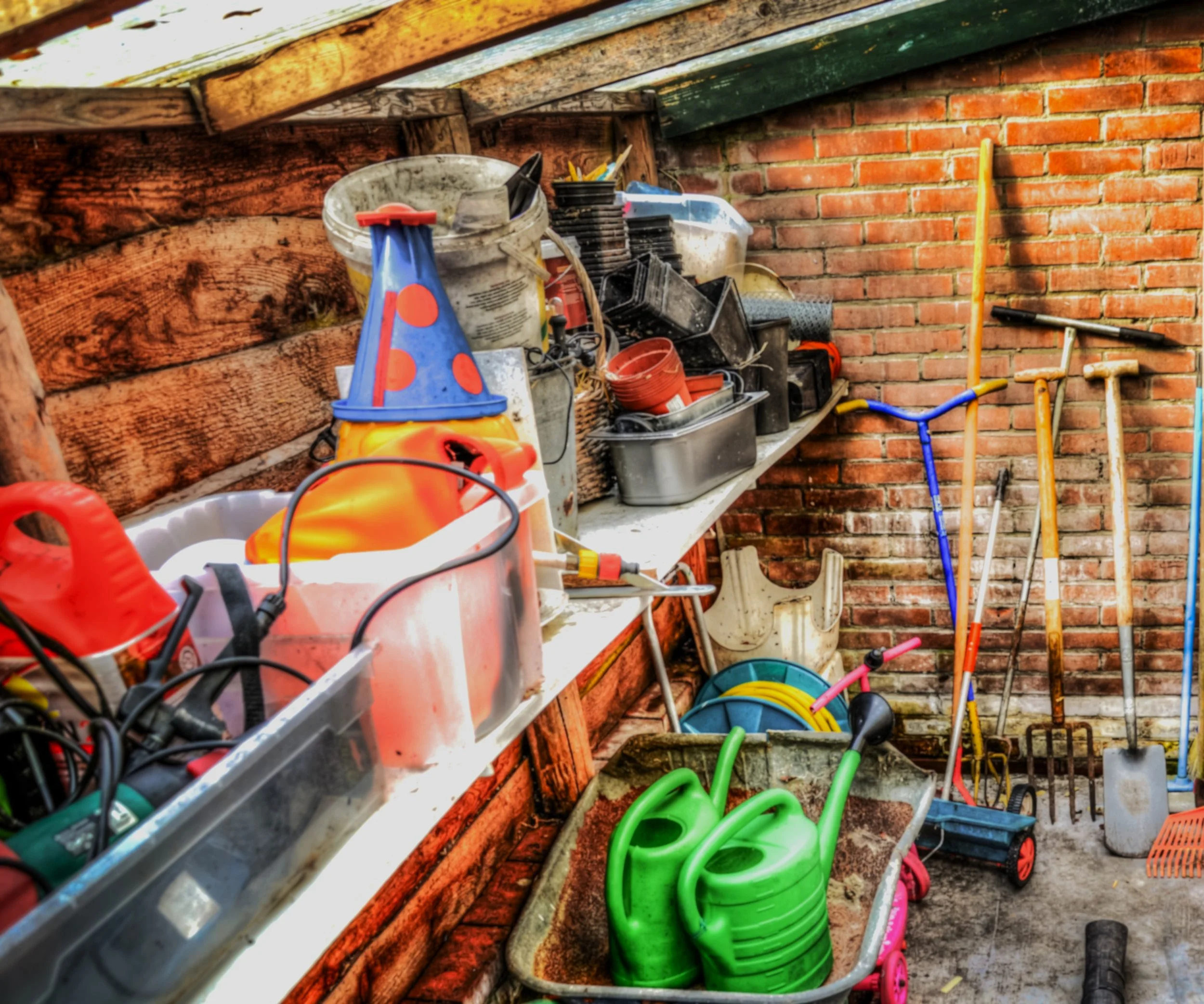 A collection of gardening and outdoor tools including a green watering can, a pink children's bicycle, and various hand tools, stored on shelves against a brick wall, in a shed or garage.