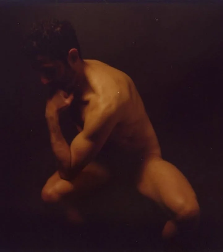 Un homme nu en pose artistique, assis avec un genou levé et la tête penchée, dans un contexte sombre.