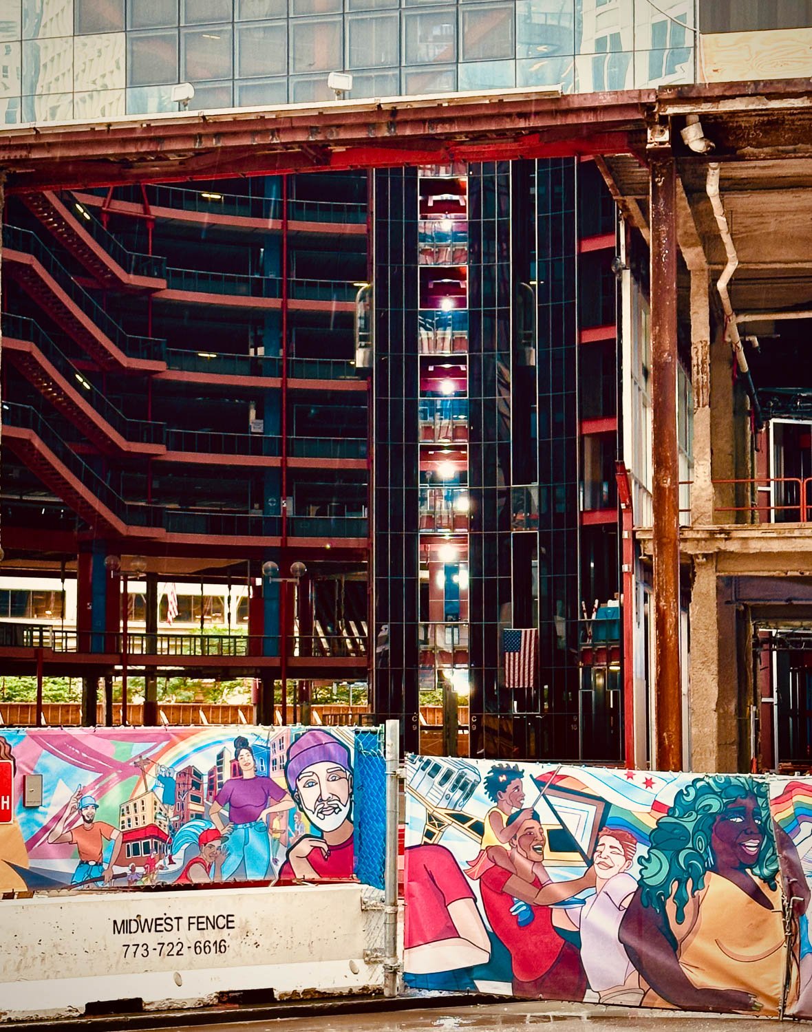 A construction site near a tall building with reflective glass windows. Colorful murals of diverse people are painted on the temporary fence surrounding the site.