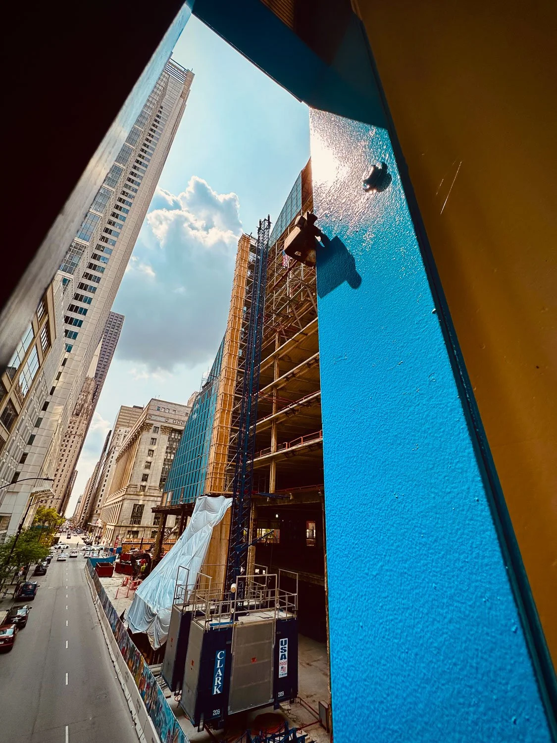 View of a construction site in a city, seen through a colorful, geometric opening. The site includes a partially built multi-story building with scaffolding and a construction crane. Tall skyscrapers and a street with moving vehicles are visible in t