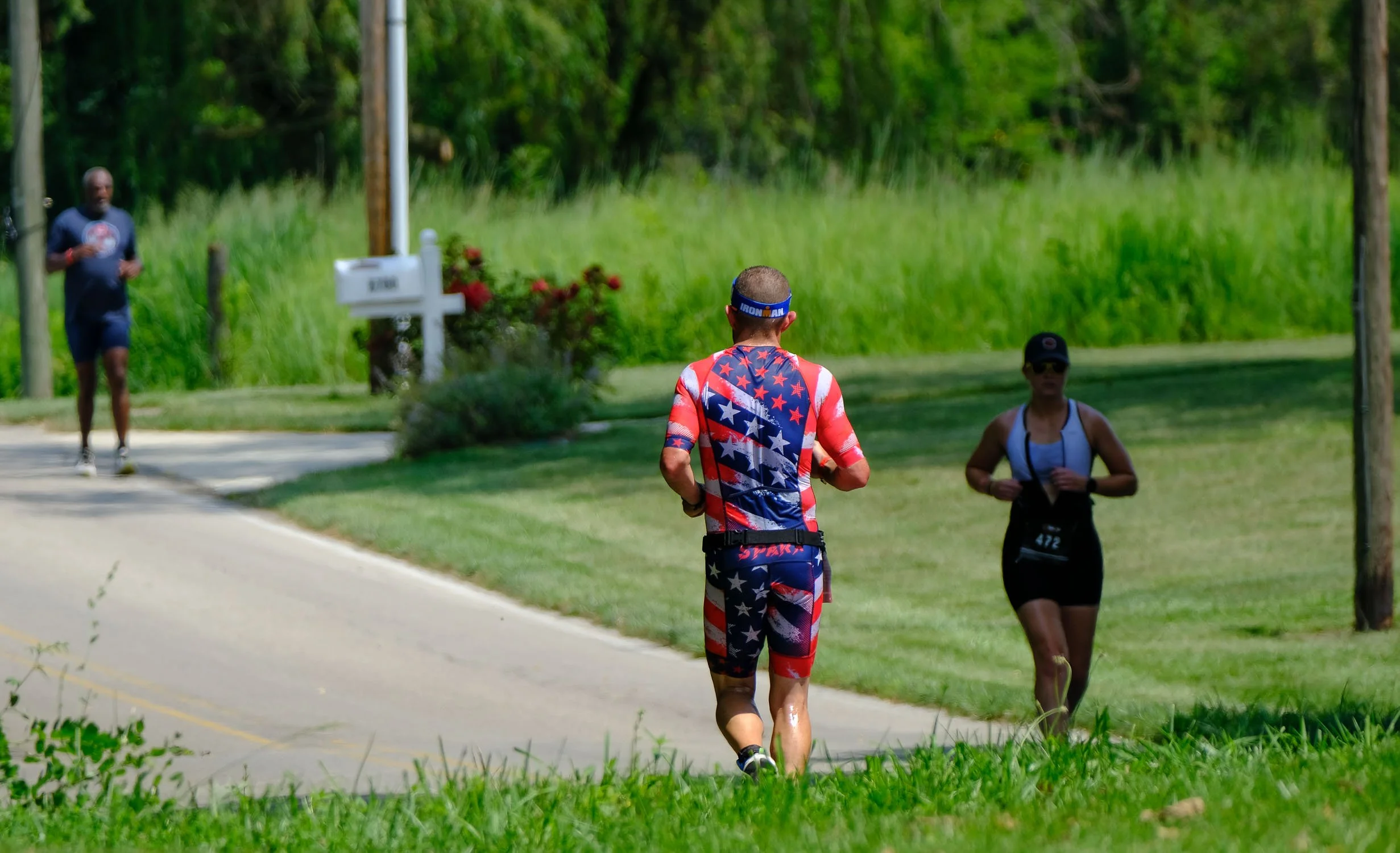 A man in patriotic running gear, decorated with stars and stripes, jogging on a rural road with two other runners in the background, surrounded by green grass and trees.