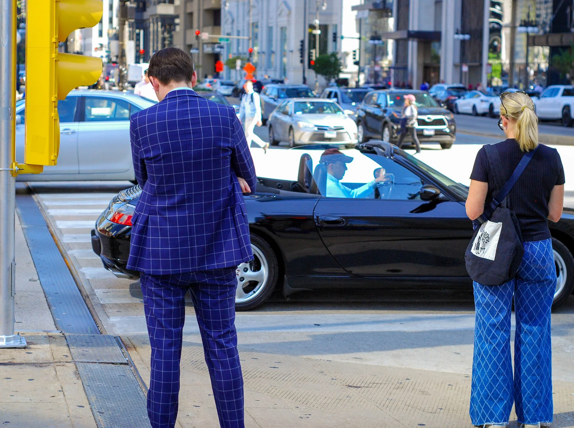 Two people standing at a crosswalk wait to cross a city street, with a black convertible car in front of them and traffic in the background.