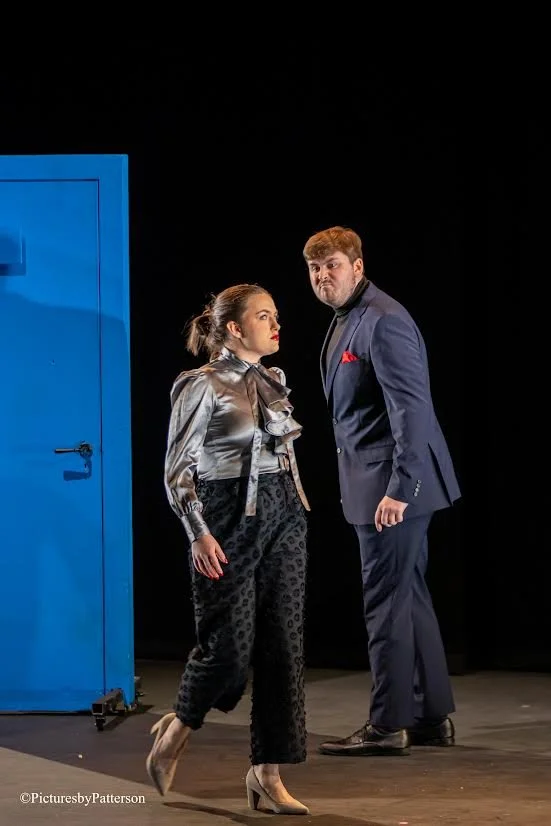 A woman in shiny silver jacket and patterned pants walking past a man in a dark suit, standing in front of a blue door on a black stage.