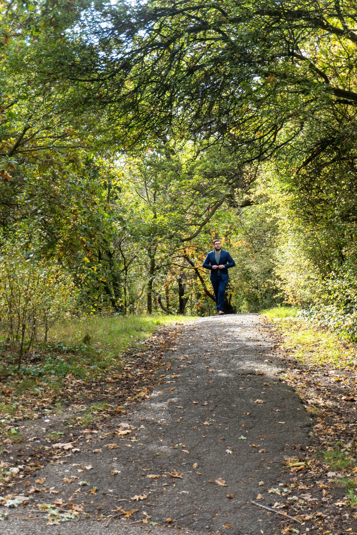 A man in a suit walking on a dirt path through a wooded area with green foliage overhead.