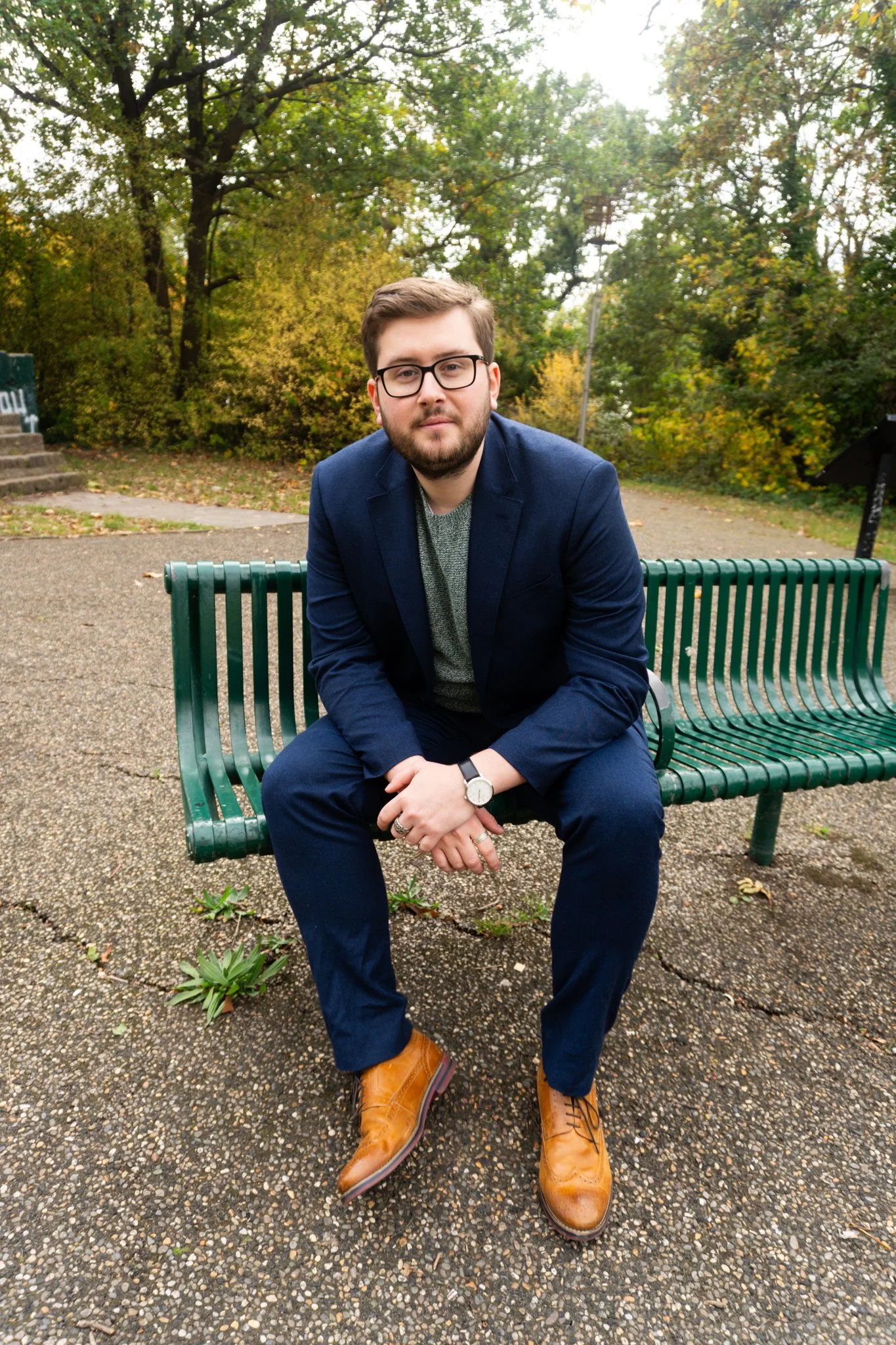 A young man with glasses, a beard, and brown hair, dressed in a navy blazer, gray shirt, navy pants, and tan boots, sitting on a green park bench in an outdoor setting with trees and bushes.