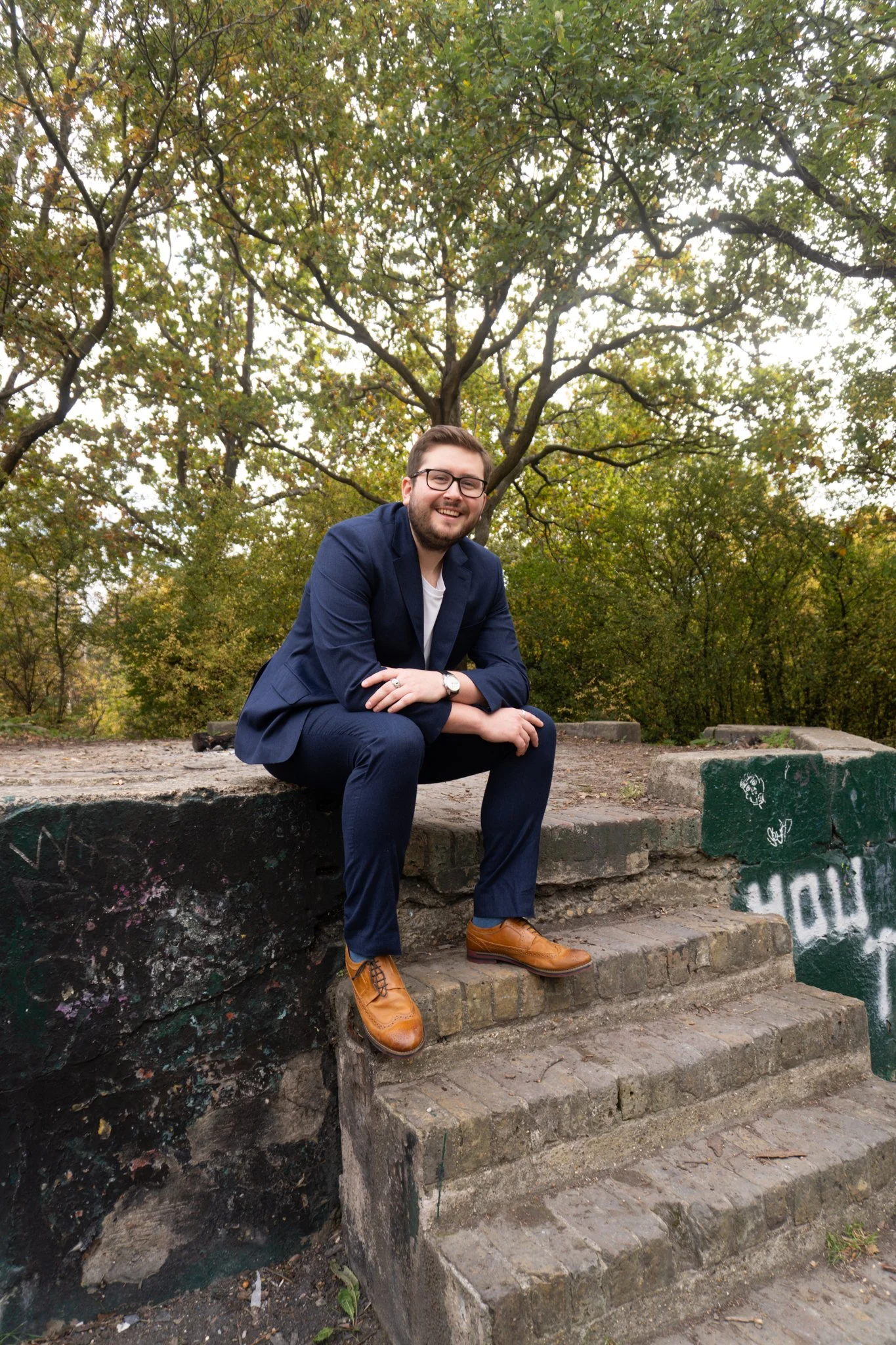 A man in a navy blue suit and brown shoes sitting on brick steps outdoors surrounded by trees with green and yellow leaves, smiling at the camera.