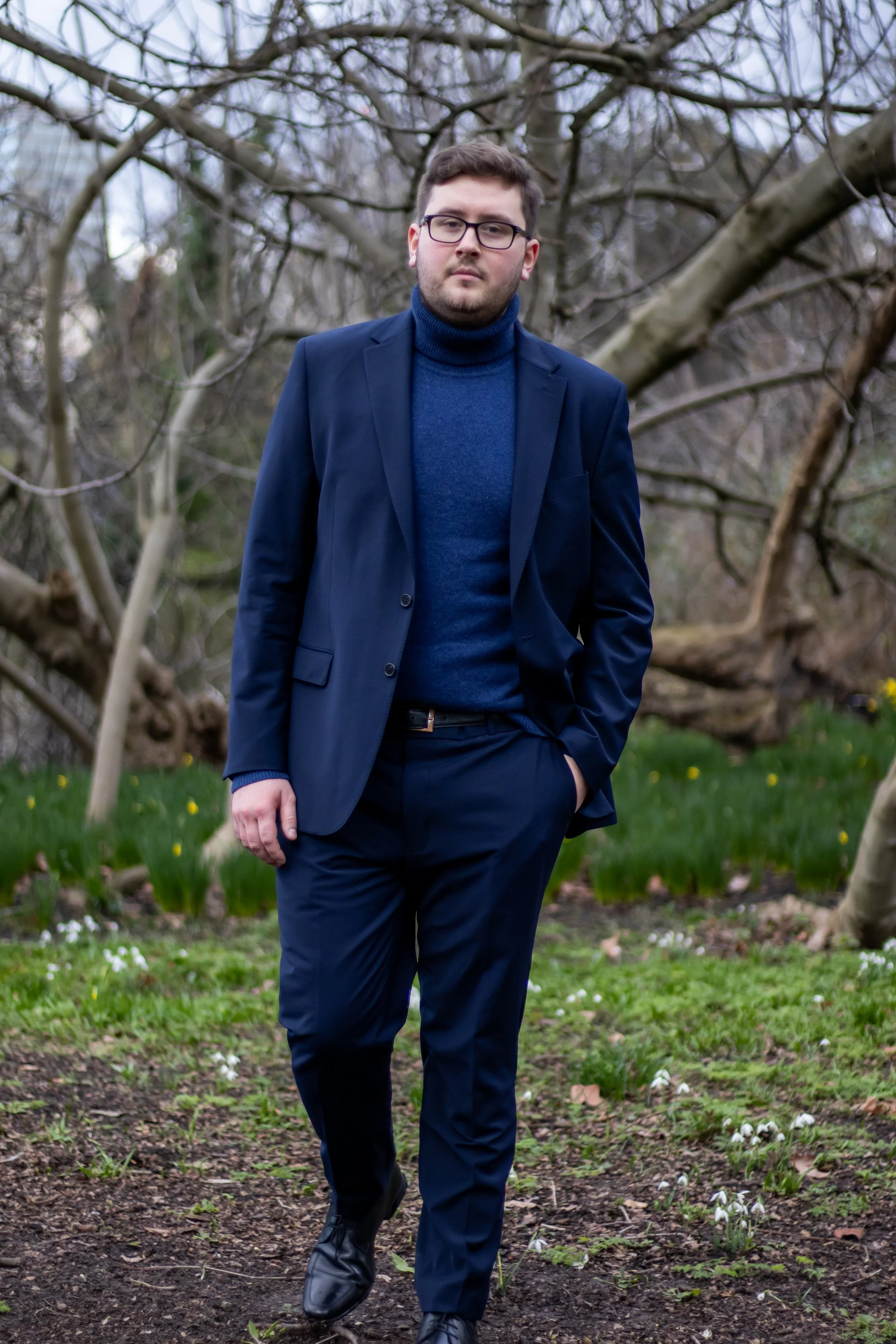 A man dressed in a navy suit with a matching turtleneck sweater, walking outdoors in a park with leafless trees and grass with small white flowers.