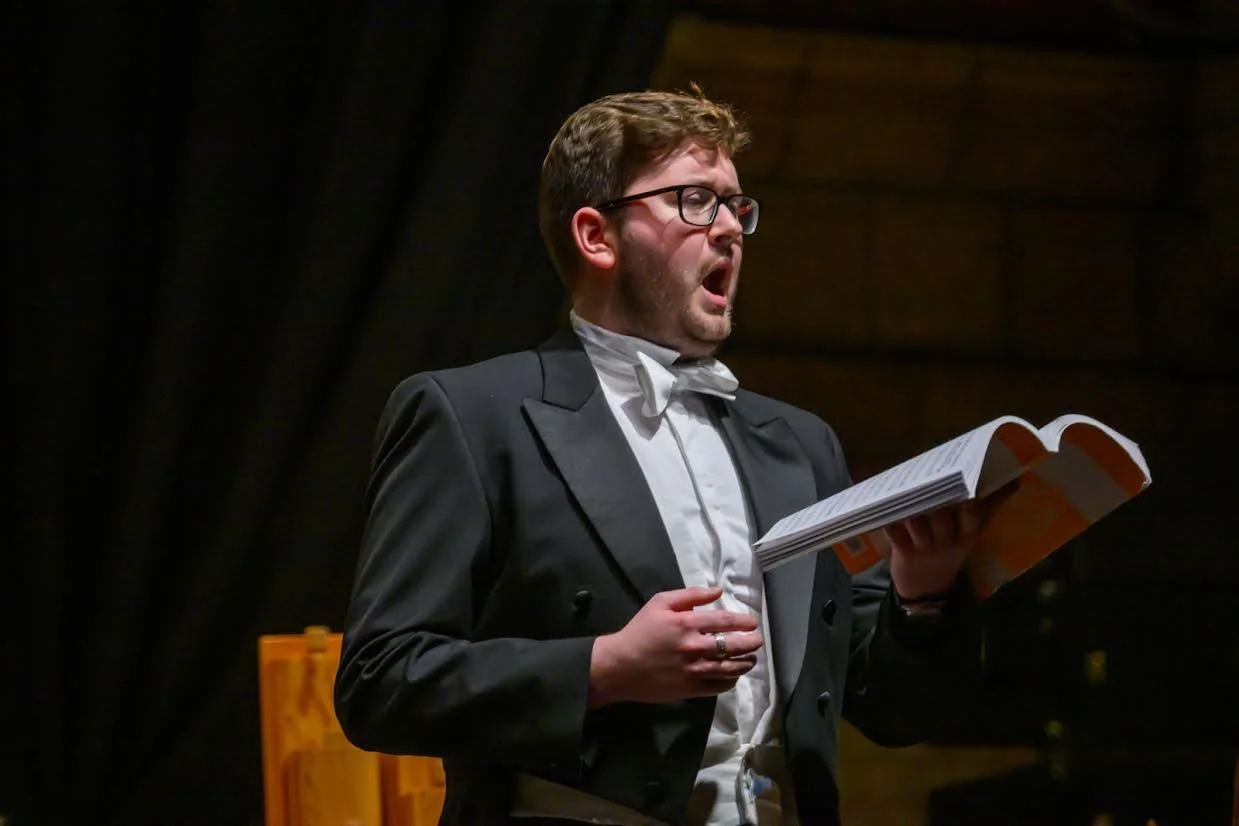 A man dressed in formal attire, including a tuxedo with a white shirt and bow tie, singing or reading from a script or sheet music in front of a dark background.