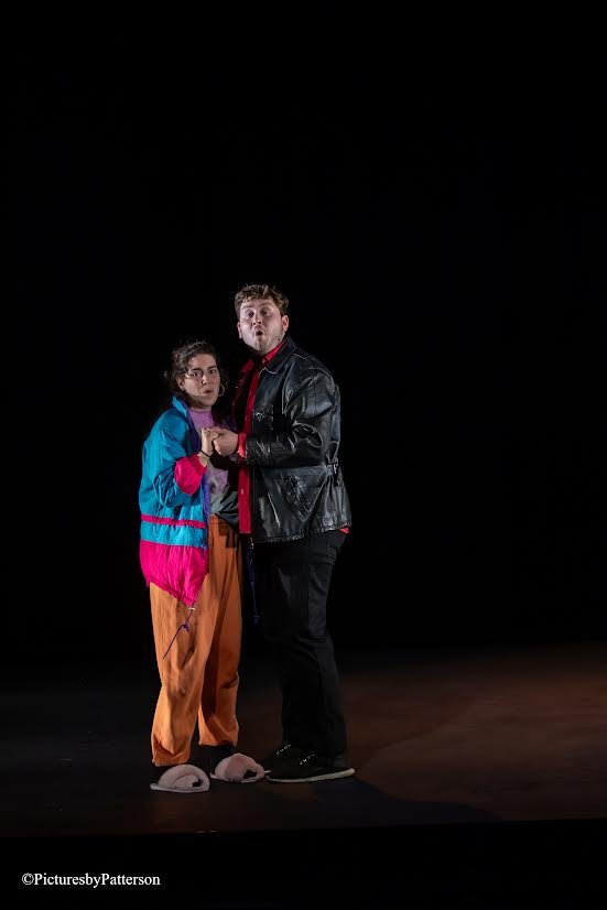 Two people standing together in a dark space, holding hands and looking surprised. The person on the left is wearing bright orange pants, a teal and pink jacket, and slippers. The person on the right is wearing a black leather jacket, black pants, an