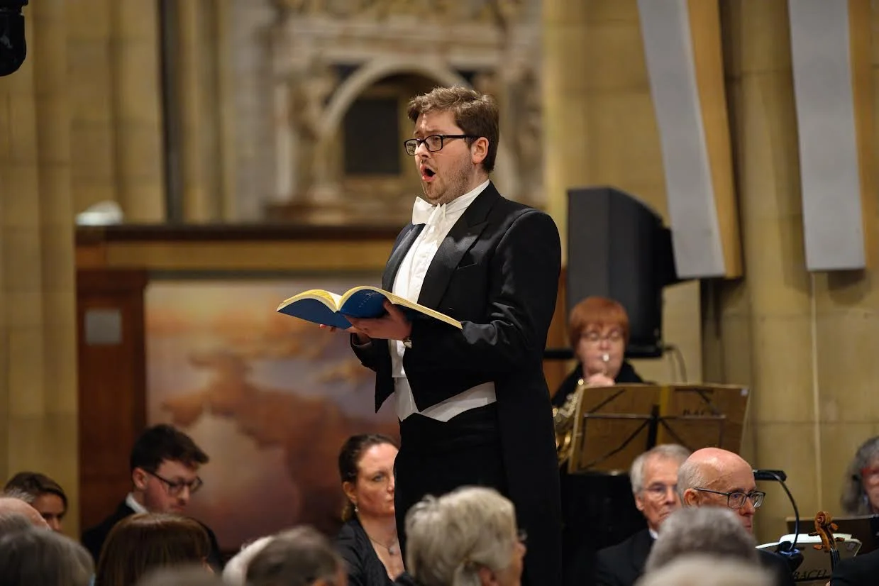 A man dressed in a tuxedo with a bow tie singing or speaking from a book in a concert hall, with an orchestra behind him and an audience watching.