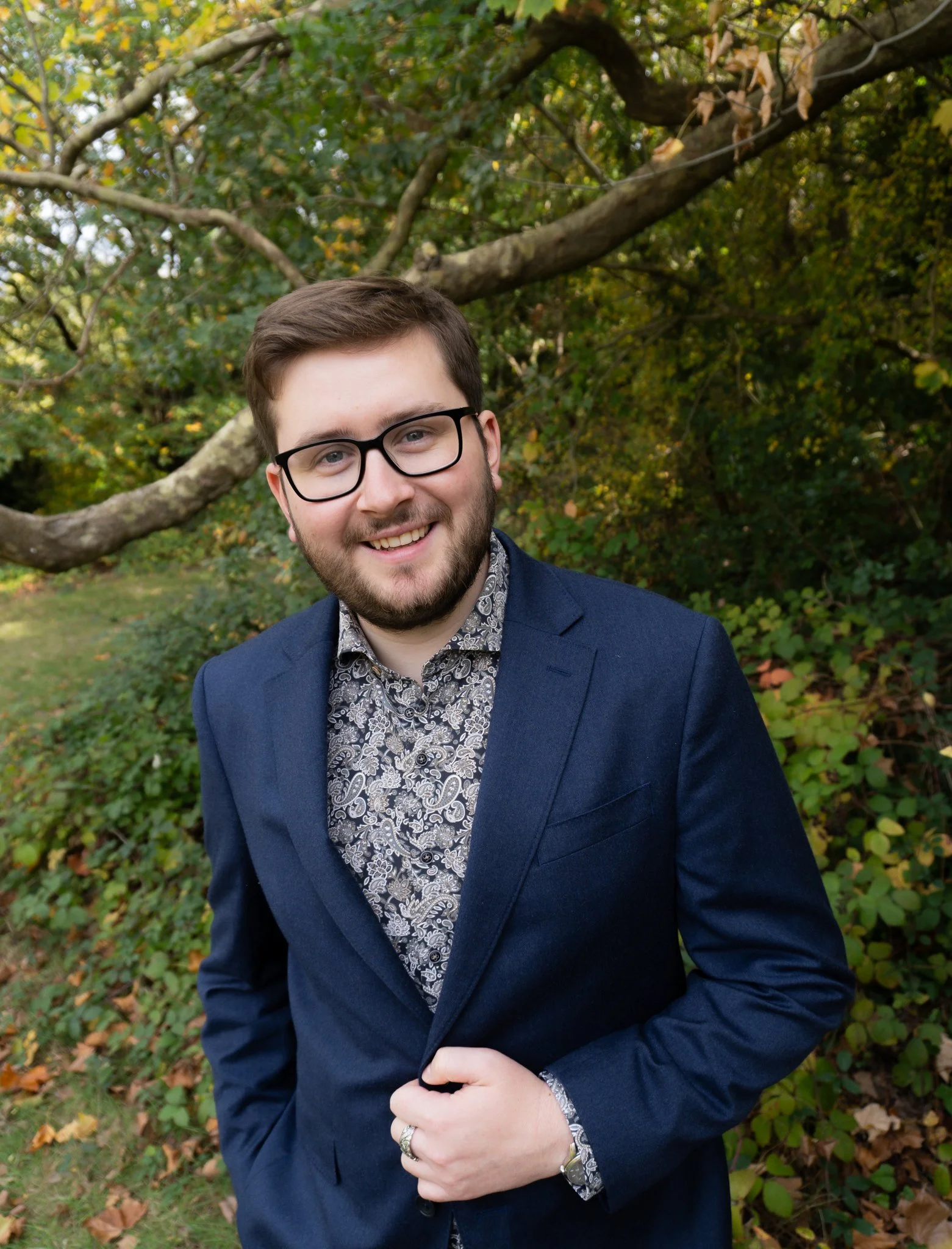 A smiling young man with glasses, wearing a patterned shirt and a dark blue blazer, standing outdoors in a park with trees and greenery in the background.