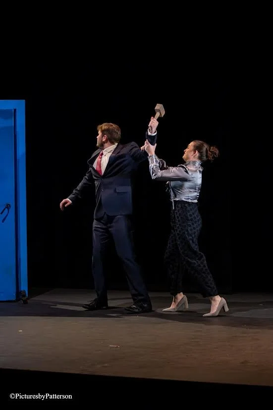 A man in a suit and a woman in stylish clothing are on stage in a theatrical performance, with the woman raising a hammer over the man, who is turning away. The scene appears tense, set against a black background.