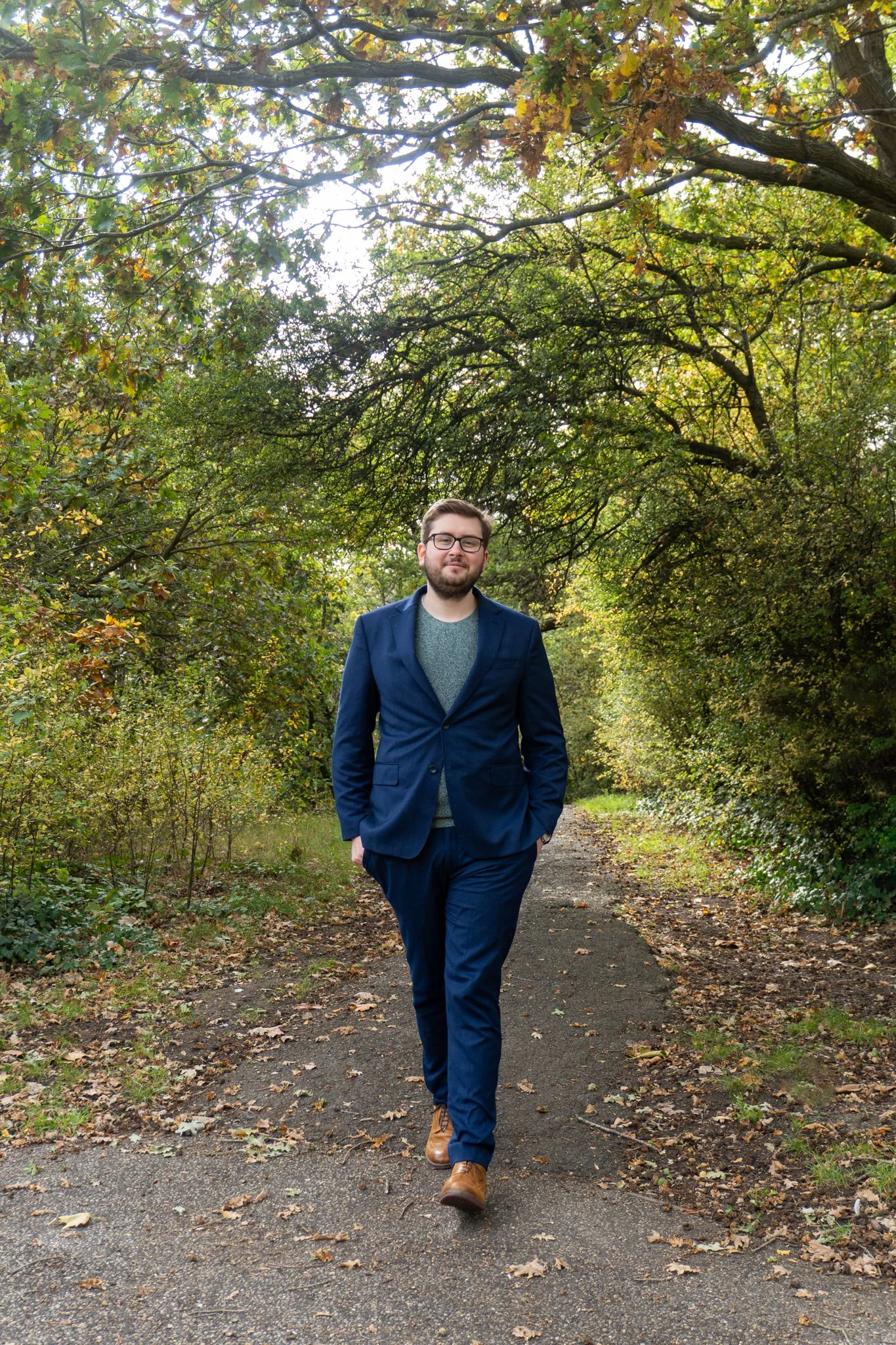 A man in a blue suit walking on a dirt path through a wooded area with fall foliage.