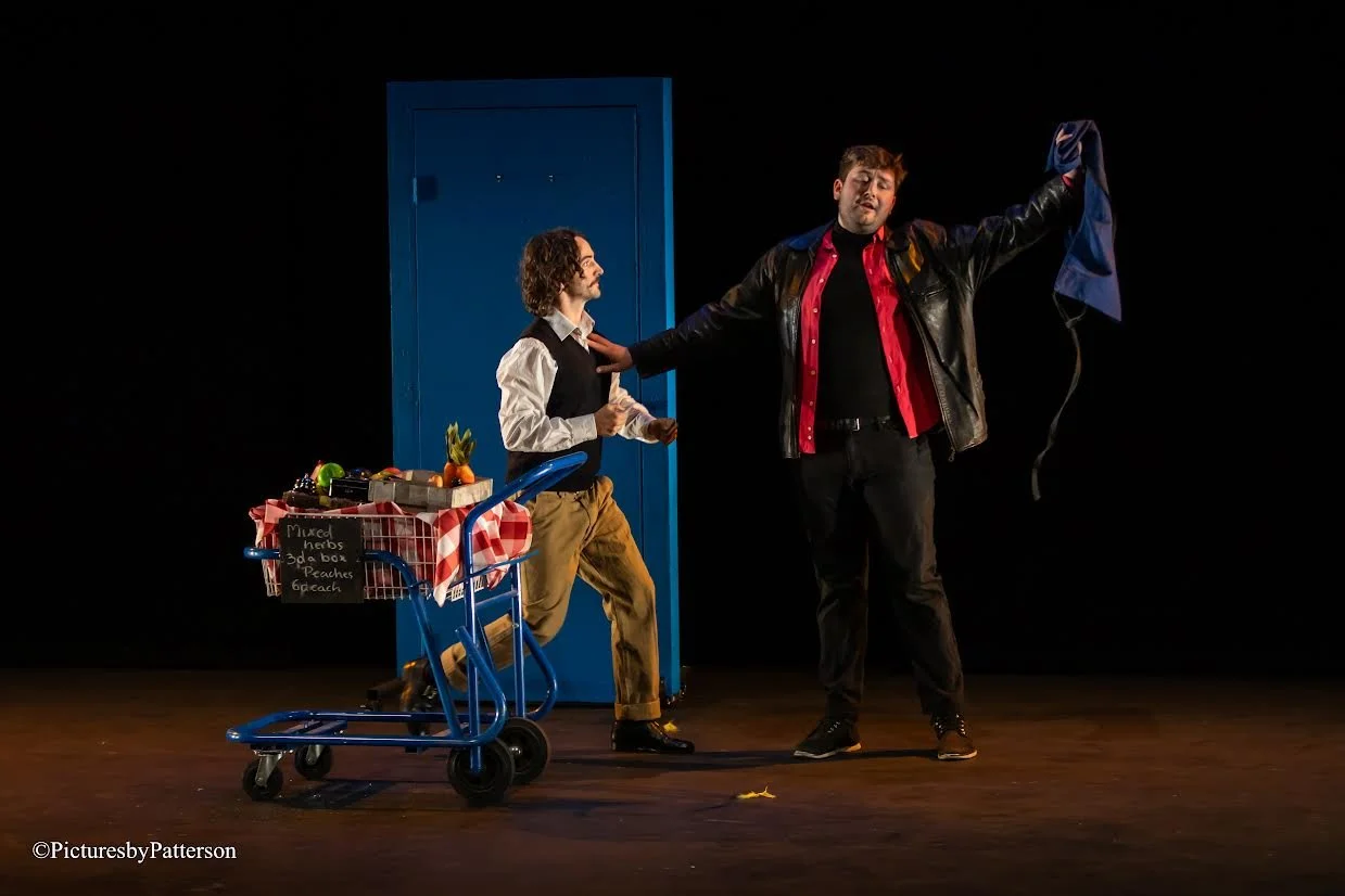 Two men on stage, one standing with a shopping cart filled with produce, and the other gesturing dramatically with his arm raised, in front of a blue door against a black background.